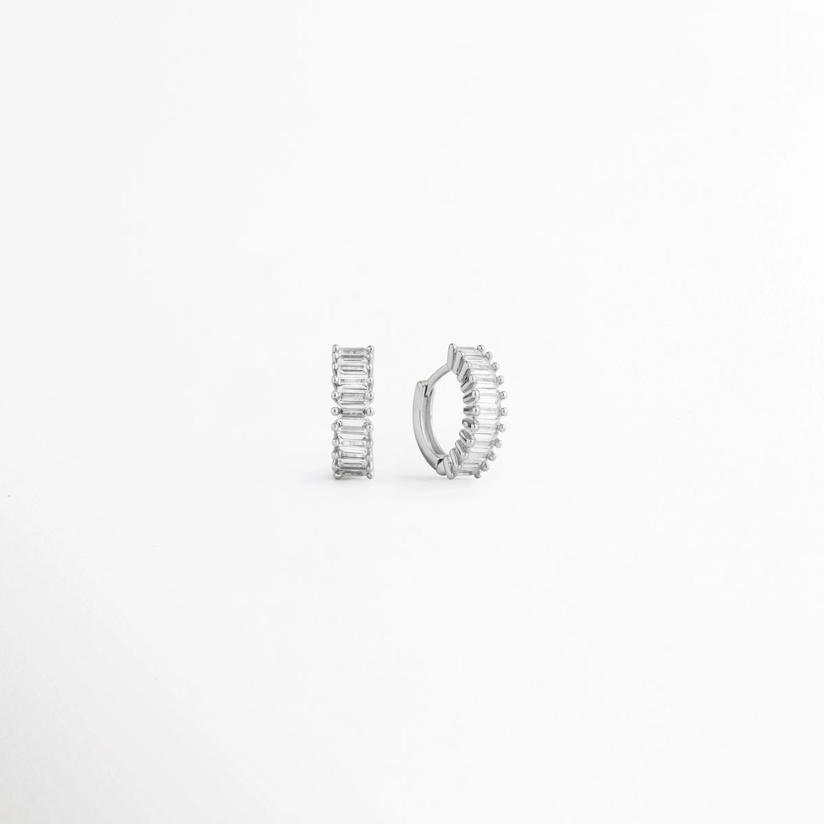 Aretes Isabel Silver Hoops mostrando perfil lateral redondo y frente con incrustaciones de circonia, Par de arrancadas pequeñas en oro blanco 18K, una de frente y otra de lado mostrando el grosor del aro, Vista angular de aretes huggies plateados Isabel Silver Hoops de 1 cm con detalle de biselado y circonias, Pendientes de aro compactos de plata y circonia, perspectiva frontal y lateral sobre fondo blanco, Aretes estilo huggie Isabel Silver Hoops cerrados en vista doble frontal y perfil.