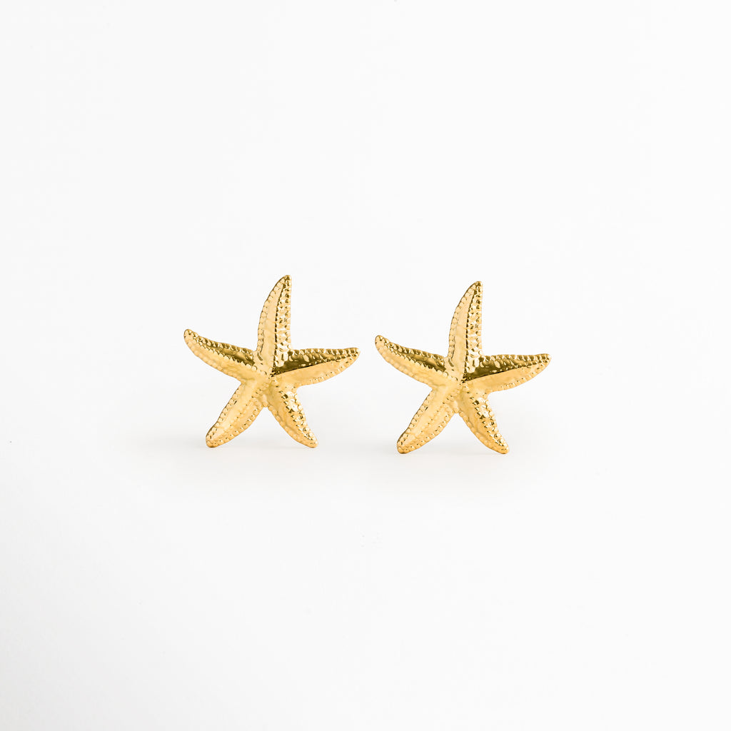 Aretes Stella en forma de estrella de mar dorada con textura rugosa y orgánica, vista frontal sobre fondo blanco, Par de topos de playa de 3.6 x 3.7 cm enchapados en oro 18K con acabado PVD en forma de estrella marina, Vista de frente de aretes marinos de acero inoxidable dorados con relieve poroso de estrella de mar, Aretes estilo stud costero en tono oro cálido con silueta realista de estrella marina sobre superficie neutra.