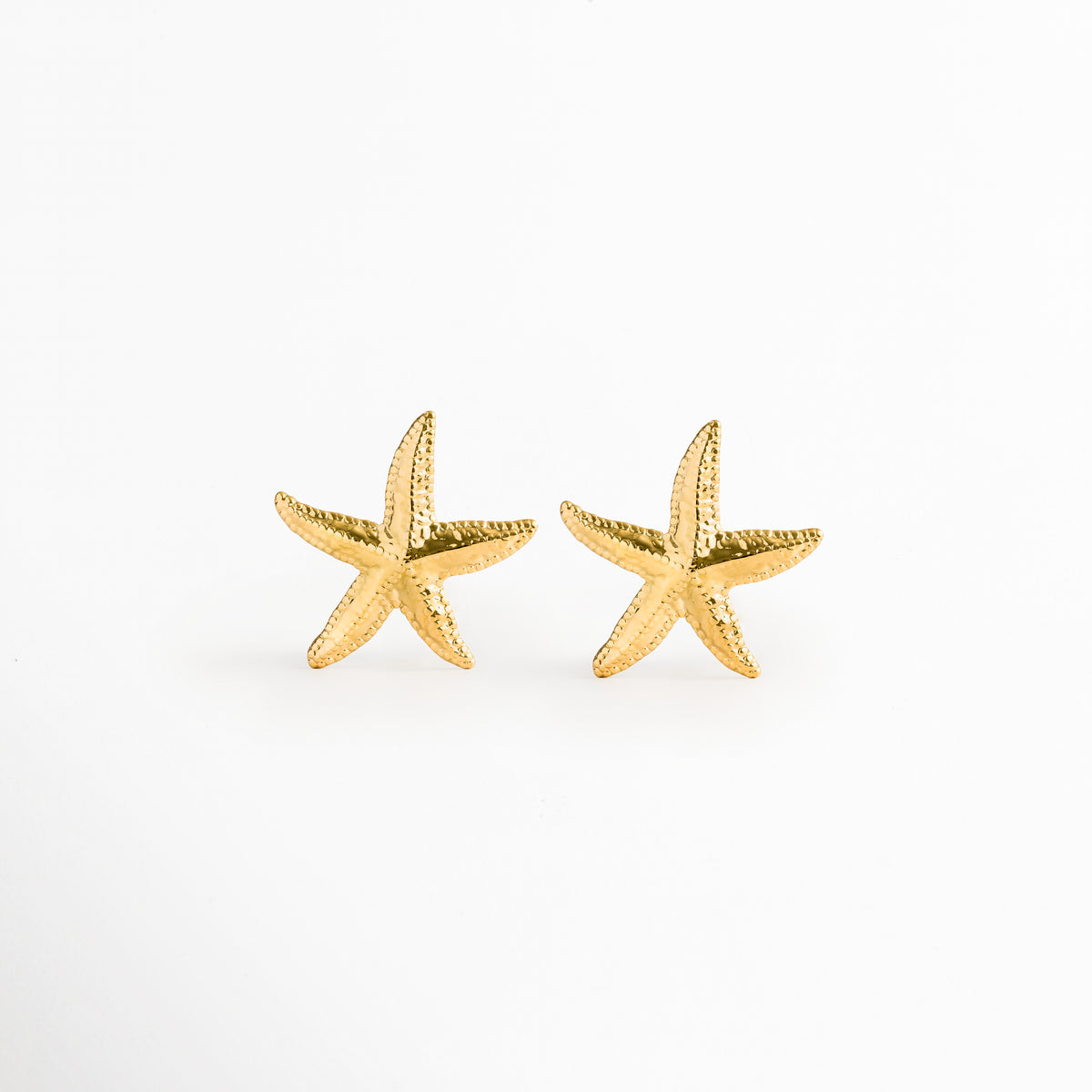 Aretes Stella en forma de estrella de mar dorada con textura rugosa y orgánica, vista frontal sobre fondo blanco, Par de topos de playa de 3.6 x 3.7 cm enchapados en oro 18K con acabado PVD en forma de estrella marina, Vista de frente de aretes marinos de acero inoxidable dorados con relieve poroso de estrella de mar, Aretes estilo stud costero en tono oro cálido con silueta realista de estrella marina sobre superficie neutra.
