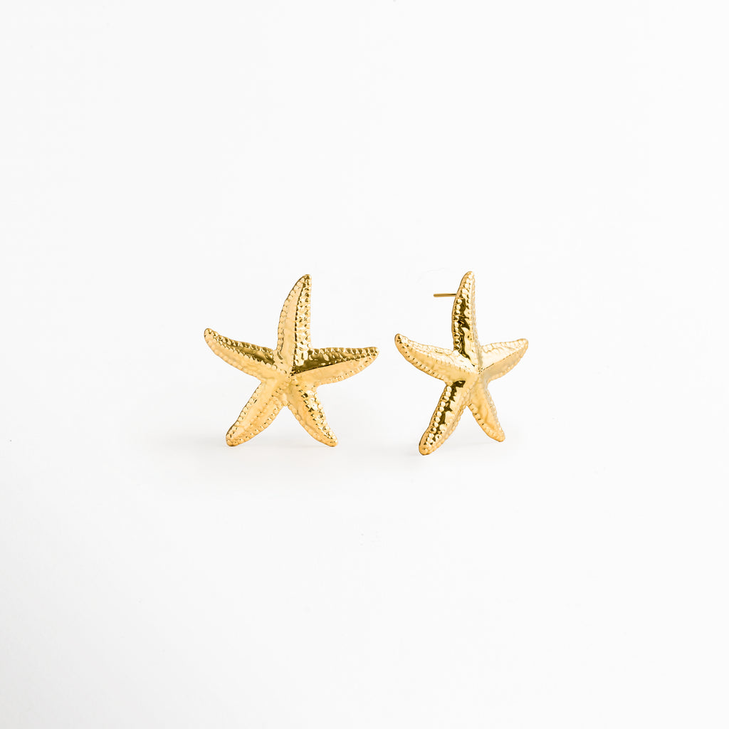 Aretes Stella dorados en perspectiva de tres cuartos evidenciando la curvatura tridimensional de los brazos de la estrella de mar, Vista inclinada de topos marinos dorados con recubrimiento PVD en oro 18K y textura orgánica, Par de aretes estivales estilo estrella de mar en toma oblicua sobre fondo claro, Detalle en ángulo del relieve poroso y los destellos dorados de los aretes Stella, Toma de tres cuartos de topos dorados de 3.7 cm de ancho con diseño