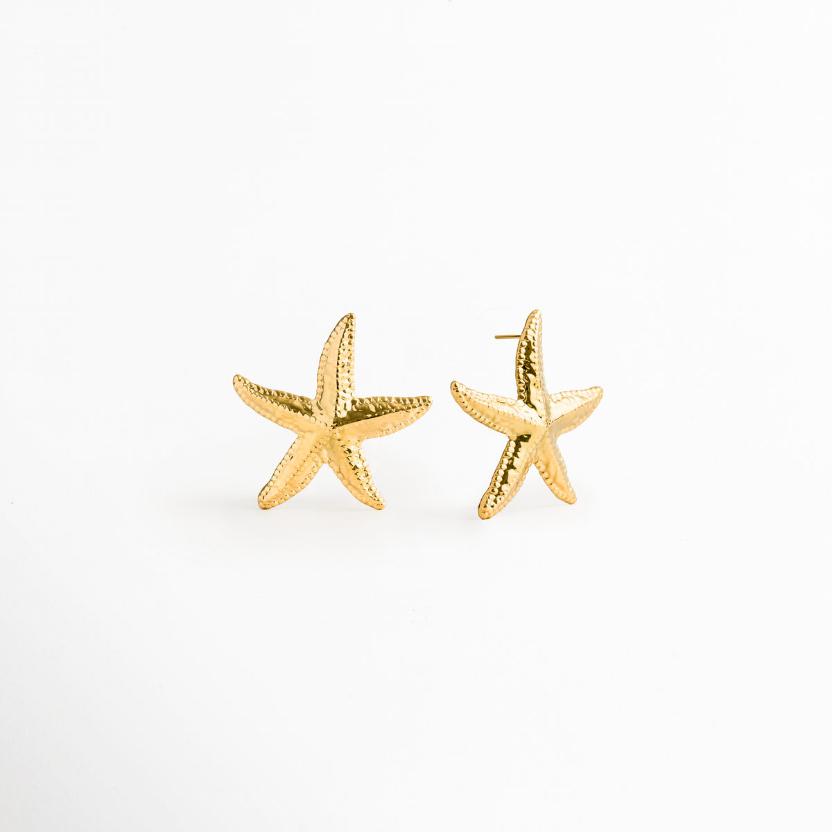 Aretes Stella dorados en perspectiva de tres cuartos evidenciando la curvatura tridimensional de los brazos de la estrella de mar, Vista inclinada de topos marinos dorados con recubrimiento PVD en oro 18K y textura orgánica, Par de aretes estivales estilo estrella de mar en toma oblicua sobre fondo claro, Detalle en ángulo del relieve poroso y los destellos dorados de los aretes Stella, Toma de tres cuartos de topos dorados de 3.7 cm de ancho con diseño