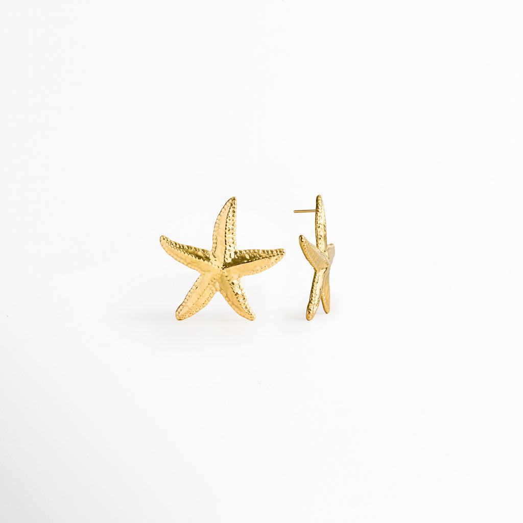 Aretes Stella en vista mixta, mostrando uno de perfil con el poste posterior recto y el otro de frente exhibiendo su silueta marina, Detalle lateral de topos dorados de estrella de mar evidenciando el grosor del metal y el poste de ajuste posterior, Vista de costado del broche de poste y curvatura de pendiente marino enchapado en oro 18K, Perspectiva de perfil de arete de acero inoxidable PVD con silueta de estrella marina y ajuste cómodo.