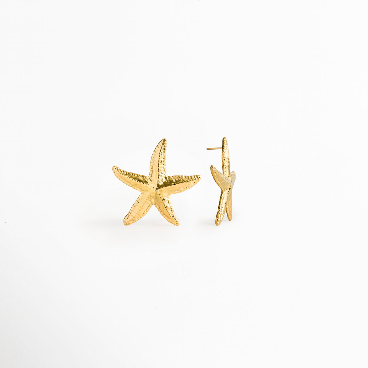 Aretes Stella en vista mixta, mostrando uno de perfil con el poste posterior recto y el otro de frente exhibiendo su silueta marina, Detalle lateral de topos dorados de estrella de mar evidenciando el grosor del metal y el poste de ajuste posterior, Vista de costado del broche de poste y curvatura de pendiente marino enchapado en oro 18K, Perspectiva de perfil de arete de acero inoxidable PVD con silueta de estrella marina y ajuste cómodo.