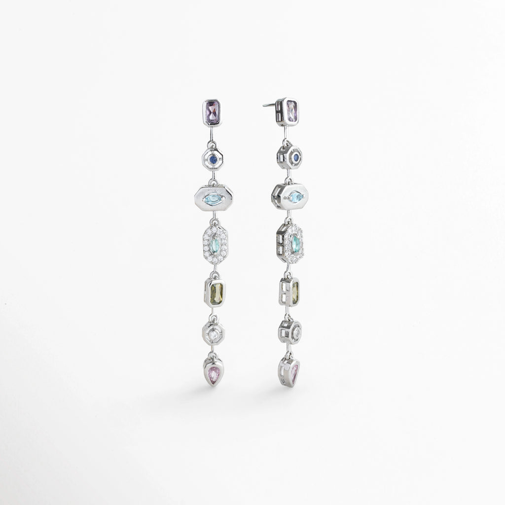 Vista en tres cuartos de los Aretes Silver Roux mostrando la fluidez y textura de la tira articulada con pedrería fina, Acercamiento en ángulo a la secuencia de gemas de colores y biseles decorativos en oro blanco 18K, Toma en perspectiva que destaca el reflejo de la luz en las gemas sintéticas multicolores de corte brillante, Muestra de la alineación estilizada y la caída fluida de 6.5 cm de los aretes colgantes sobre fondo claro.