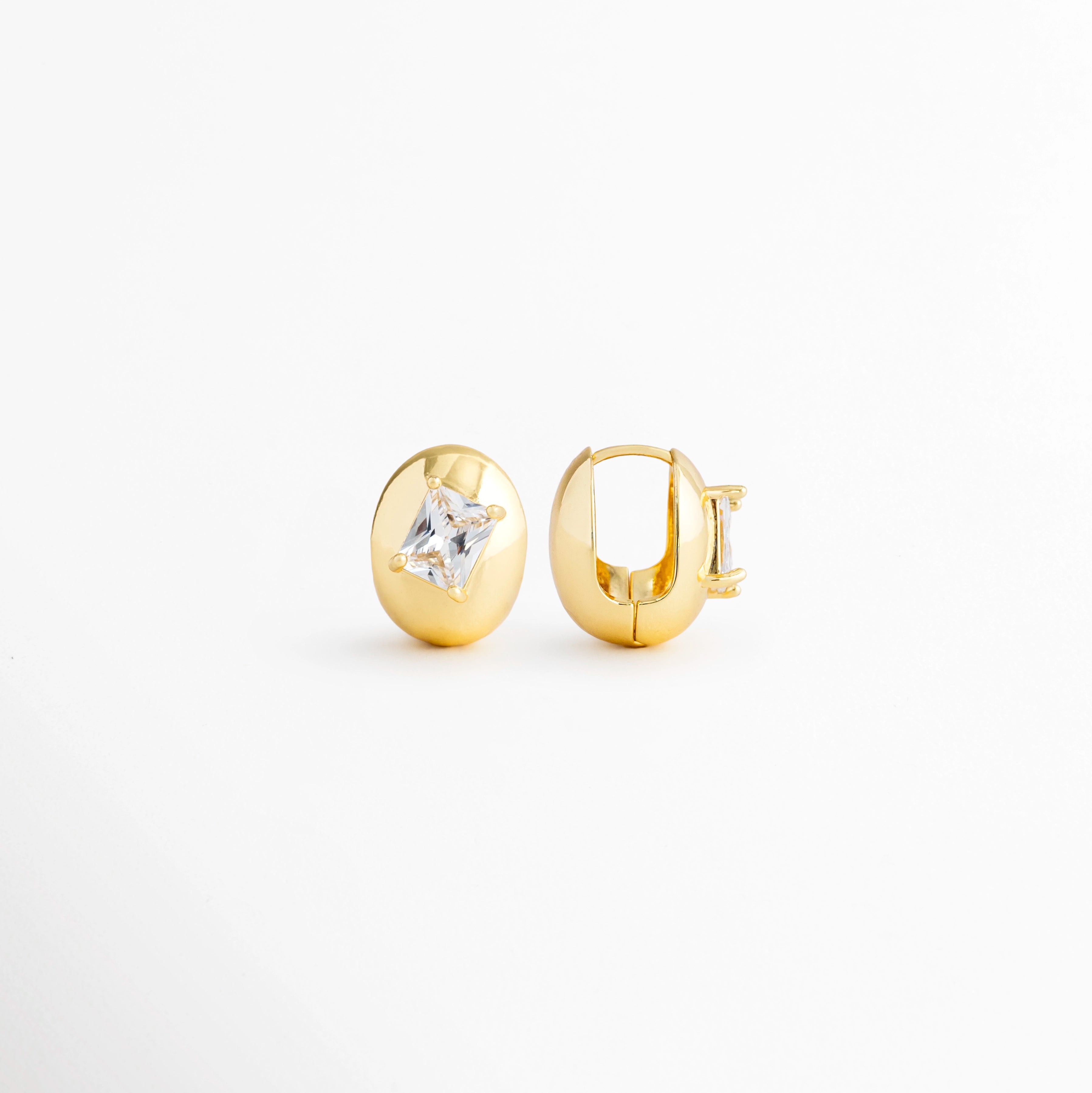 Vista en ángulo de los Aretes Piaf Hoops mostrando el perfil del aro y la zirconia central, Perspectiva de los Aretes Piaf Hoops con acabado en Oro 18K y diseño ovalado, Aretes Piaf Hoops fotografiados de perfil resaltando el brillo de la zirconia, Vista diagonal de los Aretes Piaf Hoops elaborados en cobre con recubrimiento en Oro 18K, Aretes Piaf Hoops en perspectiva mostrando su diseño contemporáneo y elegante.