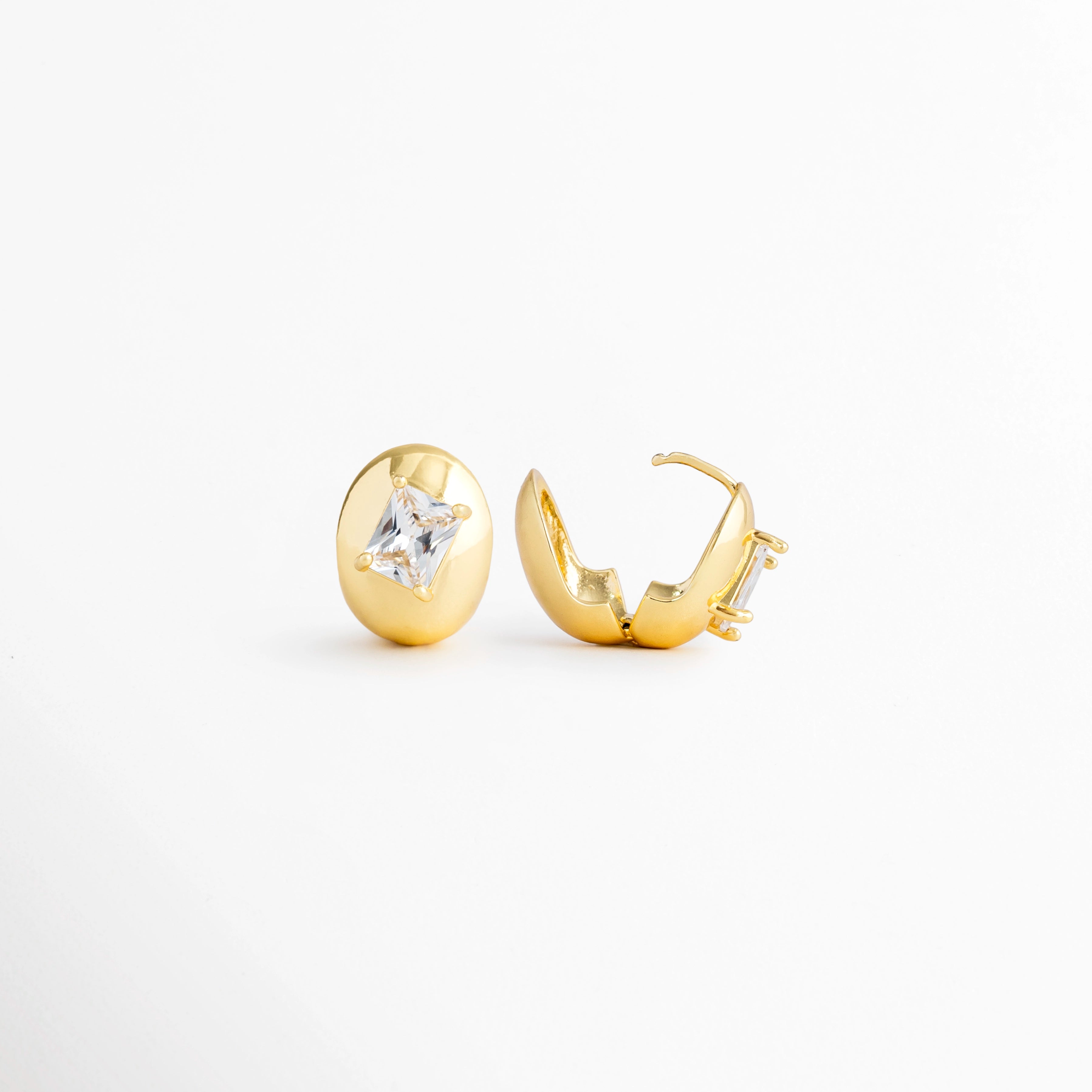 Vista lateral de los Aretes Piaf Hoops mostrando el cierre tipo bisagra y el perfil del aro, Perfil de los Aretes Piaf Hoops con acabado en Oro 18K y zirconia incrustada, Aretes Piaf Hoops en vista lateral destacando el sistema de cierre y su estructura ovalada, Detalle lateral de los Aretes Piaf Hoops elaborados en cobre con acabado dorado brillante, Vista de costado de los Aretes Piaf Hoops con diseño ovalado y zirconia central.