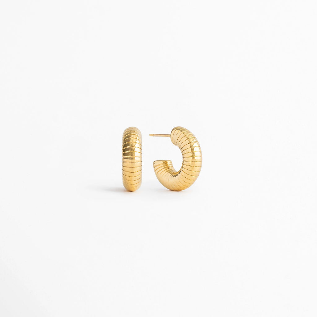 Vista en ángulo de los Aretes Golden Shell Hoops mostrando el perfil y la textura acanalada,Perspectiva de los Aretes Golden Shell Hoops con acabado en Oro 18K y diseño tipo huggie, Aretes Golden Shell Hoops fotografiados de perfil resaltando el acabado dorado brillante, Vista diagonal de los Aretes Golden Shell Hoops elaborados en acero inoxidable con recubrimiento en Oro 18K, Aretes Golden Shell Hoops en perspectiva mostrando su diseño elegante y textura acanalada.