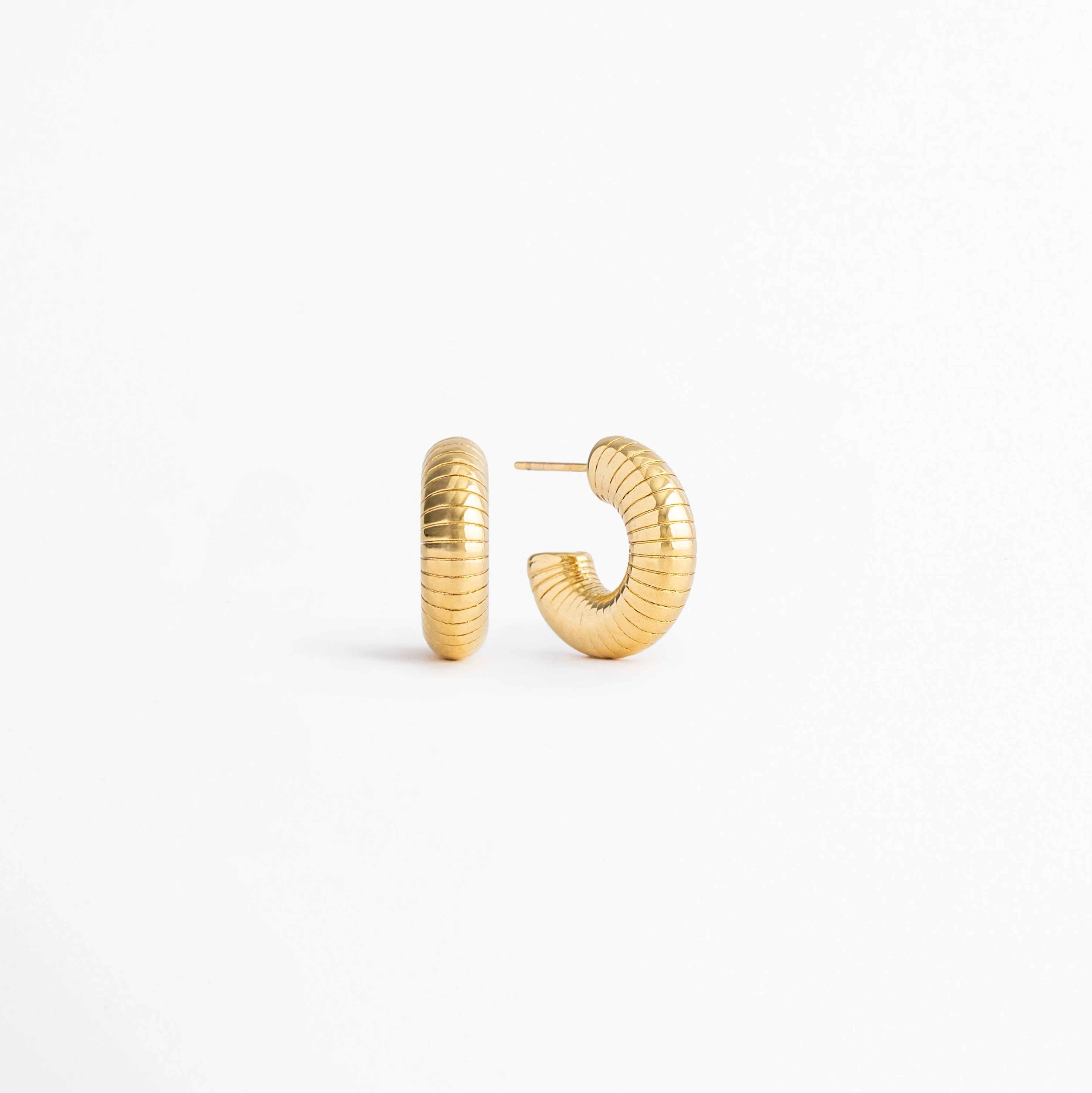 Vista en ángulo de los Aretes Golden Shell Hoops mostrando el perfil y la textura acanalada,Perspectiva de los Aretes Golden Shell Hoops con acabado en Oro 18K y diseño tipo huggie, Aretes Golden Shell Hoops fotografiados de perfil resaltando el acabado dorado brillante, Vista diagonal de los Aretes Golden Shell Hoops elaborados en acero inoxidable con recubrimiento en Oro 18K, Aretes Golden Shell Hoops en perspectiva mostrando su diseño elegante y textura acanalada.