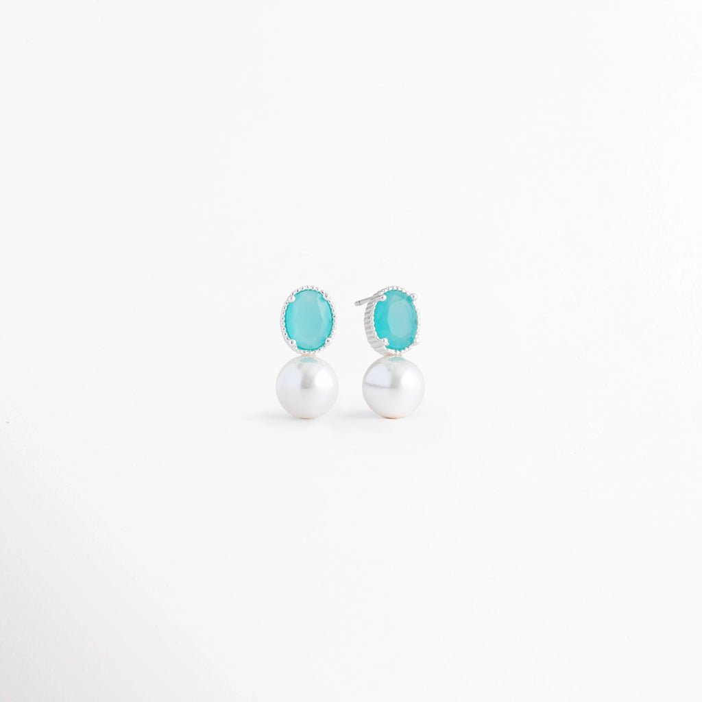 Vista en ángulo de los Aretes Boreal Blue con piedra preciosa marina azul, perla blanca y acabado en Oro Blanco 18K, Perspectiva de los Aretes Boreal Blue que resalta la piedra azul marina y la perla blanca sobre base de plata, Aretes Boreal Blue fotografiados en ángulo mostrando el brillo de la piedra marina y el acabado en Oro Blanco 18K, Vista diagonal de los Aretes Boreal Blue con diseño elegante, piedra azul y perla blanca, Aretes de plata Boreal Blue en vista inclinada con piedra preciosa marina azul 