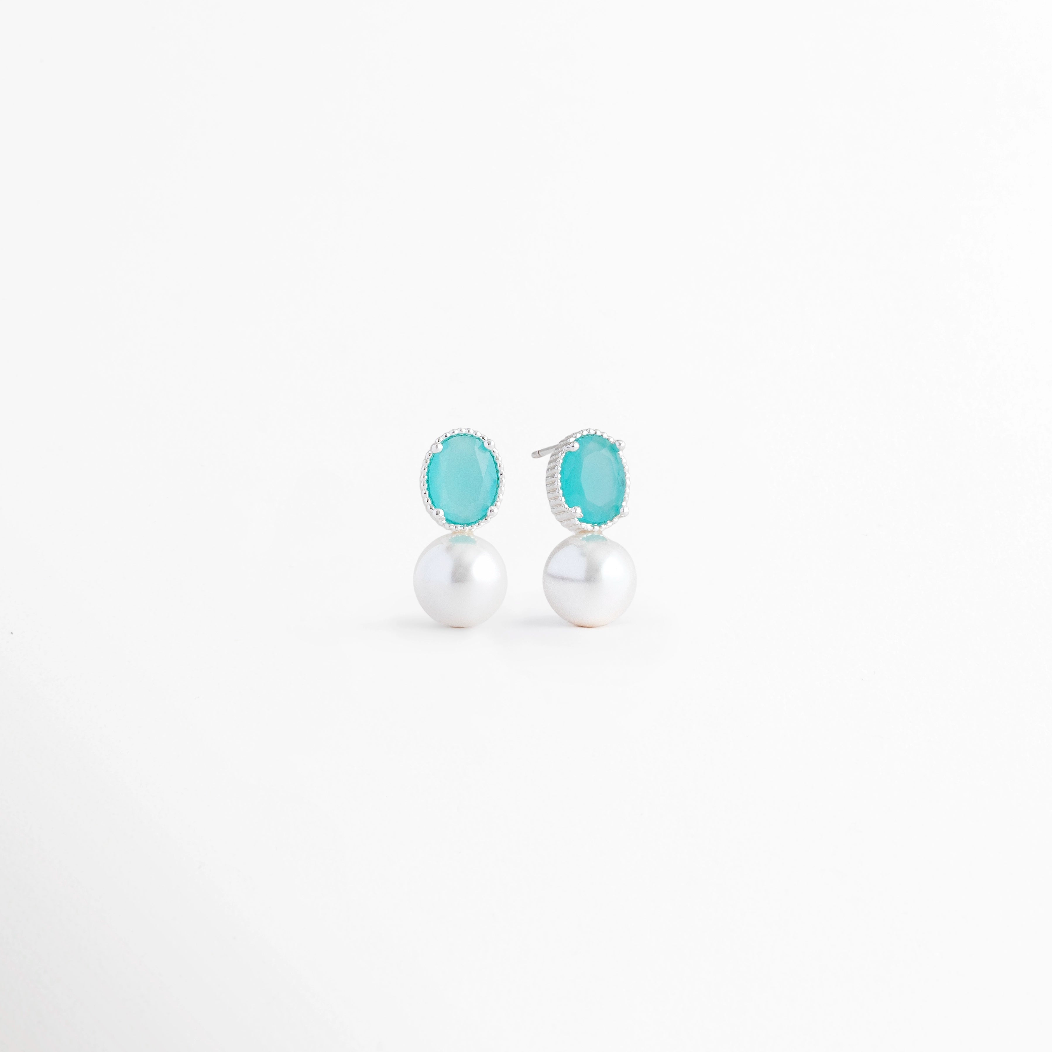 Vista en ángulo de los Aretes Boreal Blue con piedra preciosa marina azul, perla blanca y acabado en Oro Blanco 18K, Perspectiva de los Aretes Boreal Blue que resalta la piedra azul marina y la perla blanca sobre base de plata, Aretes Boreal Blue fotografiados en ángulo mostrando el brillo de la piedra marina y el acabado en Oro Blanco 18K, Vista diagonal de los Aretes Boreal Blue con diseño elegante, piedra azul y perla blanca, Aretes de plata Boreal Blue en vista inclinada con piedra preciosa marina azul 