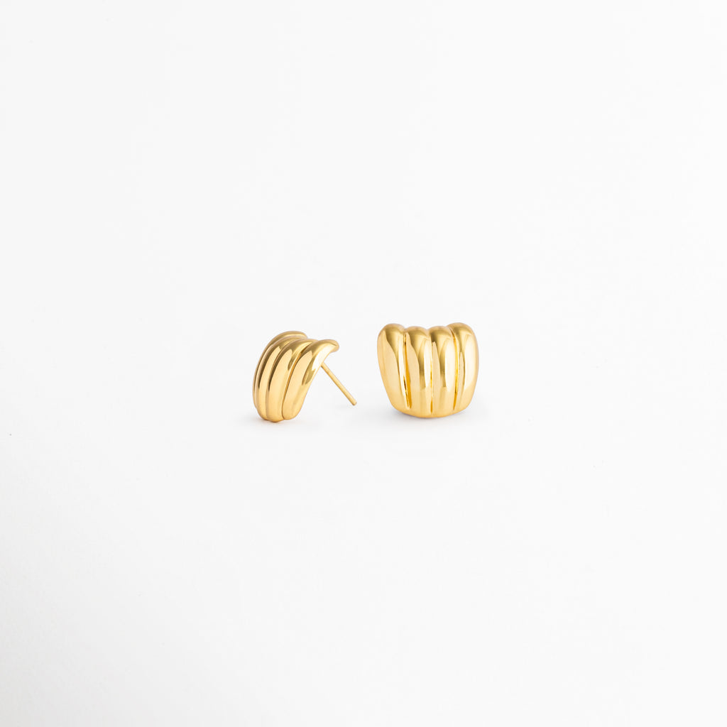 Detalle de los Aurelia Studs con acabado en oro 18K y diseño contemporáneo, Aretes dorados de acero inoxidable con textura acanalada y estilo minimalista, Vista cercana de aretes elegantes para mujer con líneas suaves y modernas, Aurelia Studs dorados, un diseño versátil para ocasiones casuales y especiales, Detalle de aretes pequeños con acabado brillante y forma curva sofisticada.