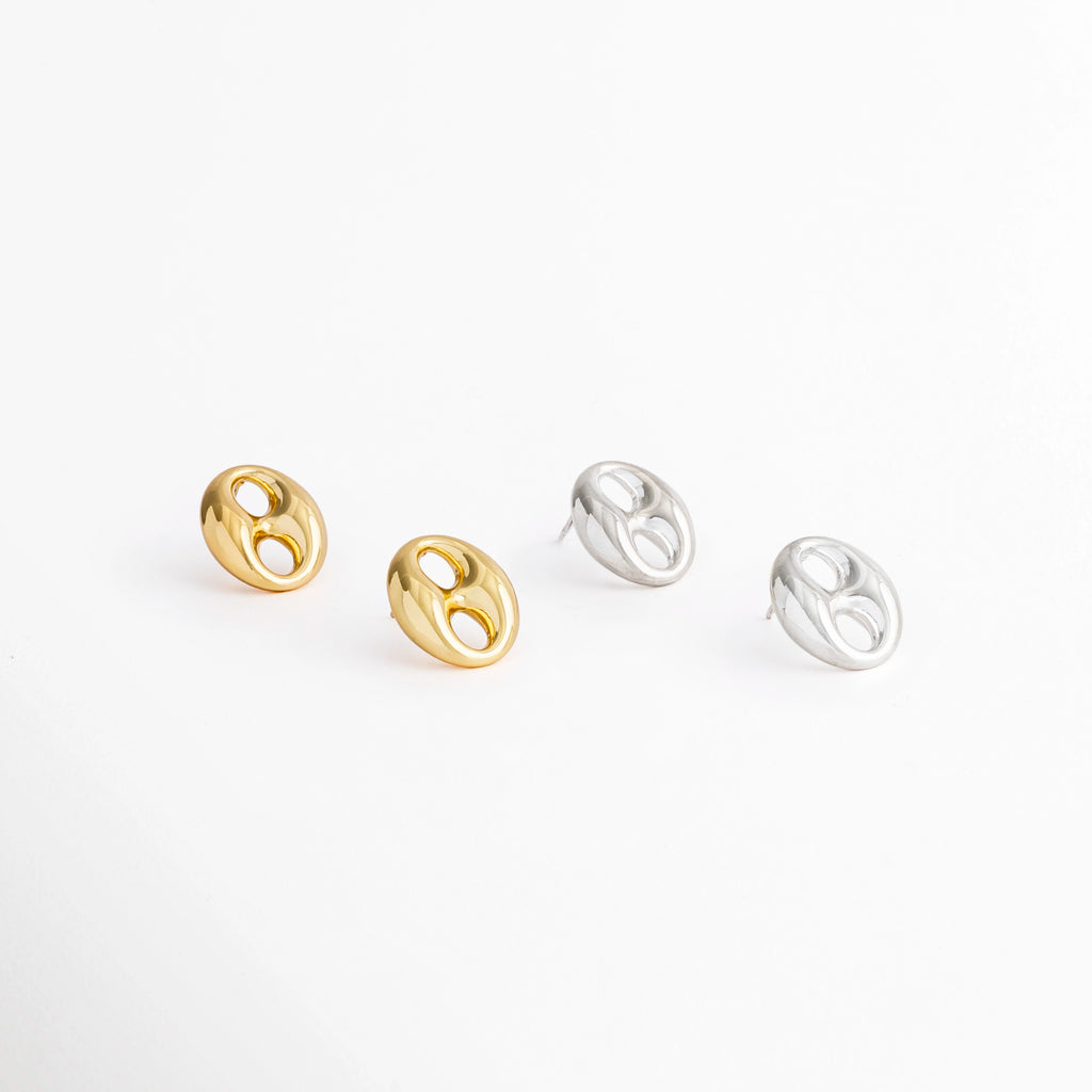 Aretes Sahra Studs disponibles en acabado Oro 18K y plateado de acero inoxidable sobre fondo blanco, Comparativa de los Aretes Sahra Studs en color dorado y plateado con diseño geométrico ovalado, Variantes de color de los Aretes Sahra Studs en acabado dorado y plateado, Aretes Sahra Studs en acabados Oro 18K y plateado mostrando ambas opciones de color, Vista comparativa de los Aretes Sahra Studs disponibles en dorado y plateado.
