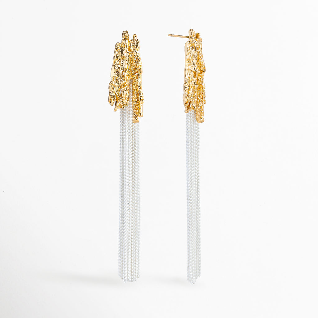 Vista de perfil de los Aretes Grutas mostrando el poste de sujeción para oreja perforada y la textura tridimensional del tope dorado, Acercamiento lateral a los aretes colgantes resaltando la estructura rocosa enchapada en oro 18K y la caída vertical de las cadenas, Toma en ángulo que muestra la profundidad y el volumen de la textura escultural inspirada en las estalactitas de cueva, Muestra de la proporción de 8.7 cm de largo de los aretes y el acabado pulido bitono.
