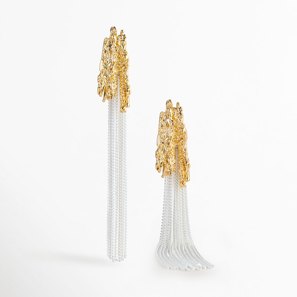 Muestra en movimiento de un arete Grutas extendido con la cascada de cadenas plateadas abriéndose suavemente, Acercamiento al contraste entre el poste rígido dorado de textura rústica y la fluidez del manojo de cadenas en tono oro blanco, Vista detallada de la caída ligera de los flecos de cadena de 8.7 cm de longitud, Plano en perspectiva que resalta los destellos de luz sobre la superficie martillada y las cadenas finas.