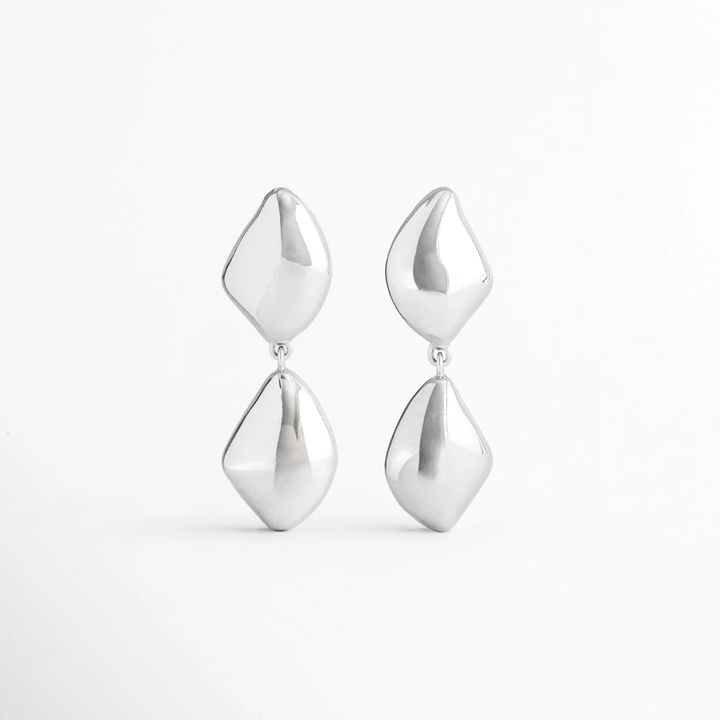 Aretes Bastet de aleación de metales con acabado plateado brillante y diseño geométrico colgante sobre fondo blanco, Vista frontal de los Aretes Bastet con figuras geométricas de acabado pulido y estilo contemporáneo, Aretes Bastet con diseño colgante de formas geométricas y acabado electro-chapado brillante, Par de Aretes Bastet con diseño moderno de doble pieza geométrica en color plateado, Aretes Bastet de estilo contemporáneo con acabado brillante y diseño geométrico alargado
