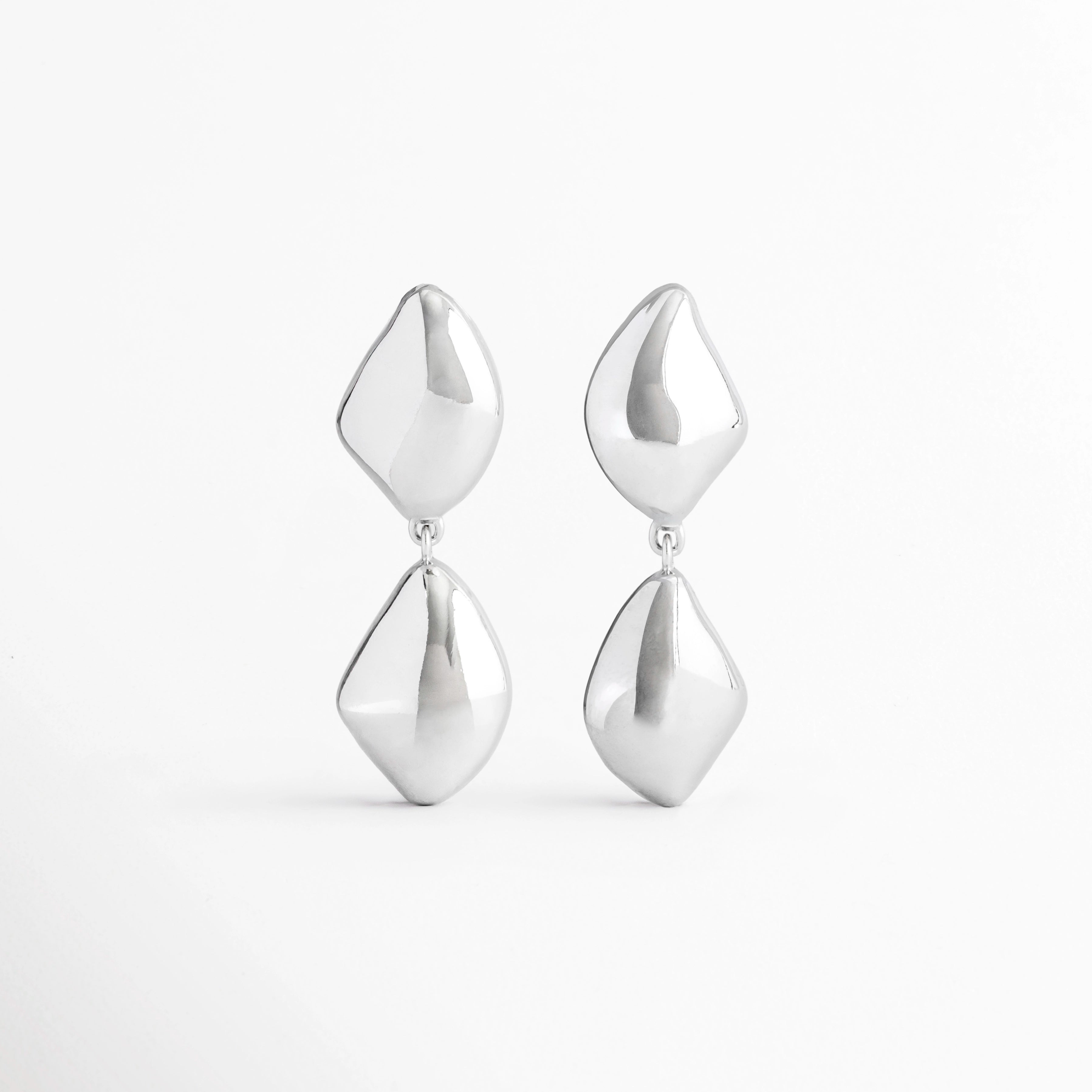 Aretes Bastet de aleación de metales con acabado plateado brillante y diseño geométrico colgante sobre fondo blanco, Vista frontal de los Aretes Bastet con figuras geométricas de acabado pulido y estilo contemporáneo, Aretes Bastet con diseño colgante de formas geométricas y acabado electro-chapado brillante, Par de Aretes Bastet con diseño moderno de doble pieza geométrica en color plateado, Aretes Bastet de estilo contemporáneo con acabado brillante y diseño geométrico alargado