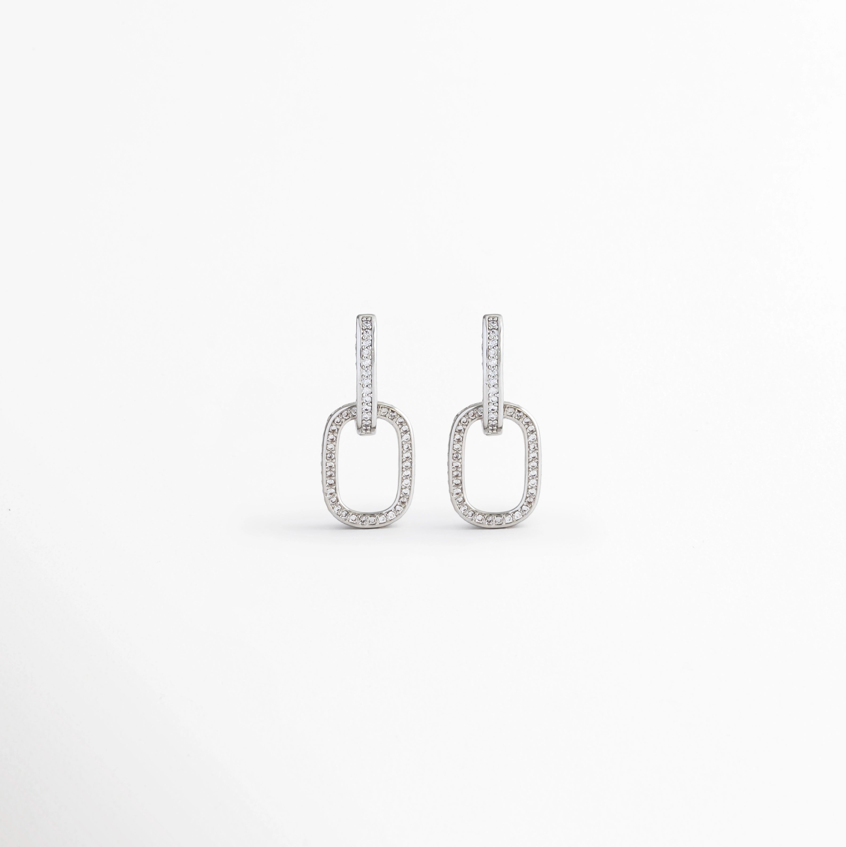 Aretes Silver Oria de cobre con recubrimiento en Oro Blanco 14K e incrustación de zirconia sobre fondo blanco, Vista frontal de los Aretes Silver Oria con diseño rectangular y zirconia en acabado de Oro Blanco 14K, Aretes Silver Oria con diseño geométrico de eslabón rectangular y zirconia brillante, Par de Aretes Silver Oria con acabado plateado y diseño minimalista para mujer, Aretes Silver Oria con forma rectangular y acabado en Oro Blanco 14K.