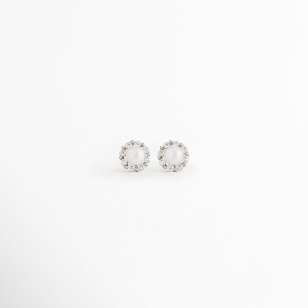 Aretes Zerina Silver Studs de Plata de Ley 925 con recubrimiento en Oro Blanco 14K y zirconia brillante sobre fondo blanco, Vista frontal de los Aretes Zerina Silver Studs con zirconia central rodeada por un delicado halo brillante, Aretes Zerina Silver Studs con diseño redondo y zirconia en acabado de Oro Blanco 14K, Par de Aretes Zerina Silver Studs elaborados en Plata de Ley 925 con zirconia y acabado plateado brillante, Aretes plateados Zerina Silver Studs con zirconia en diseño clásico y elegante.