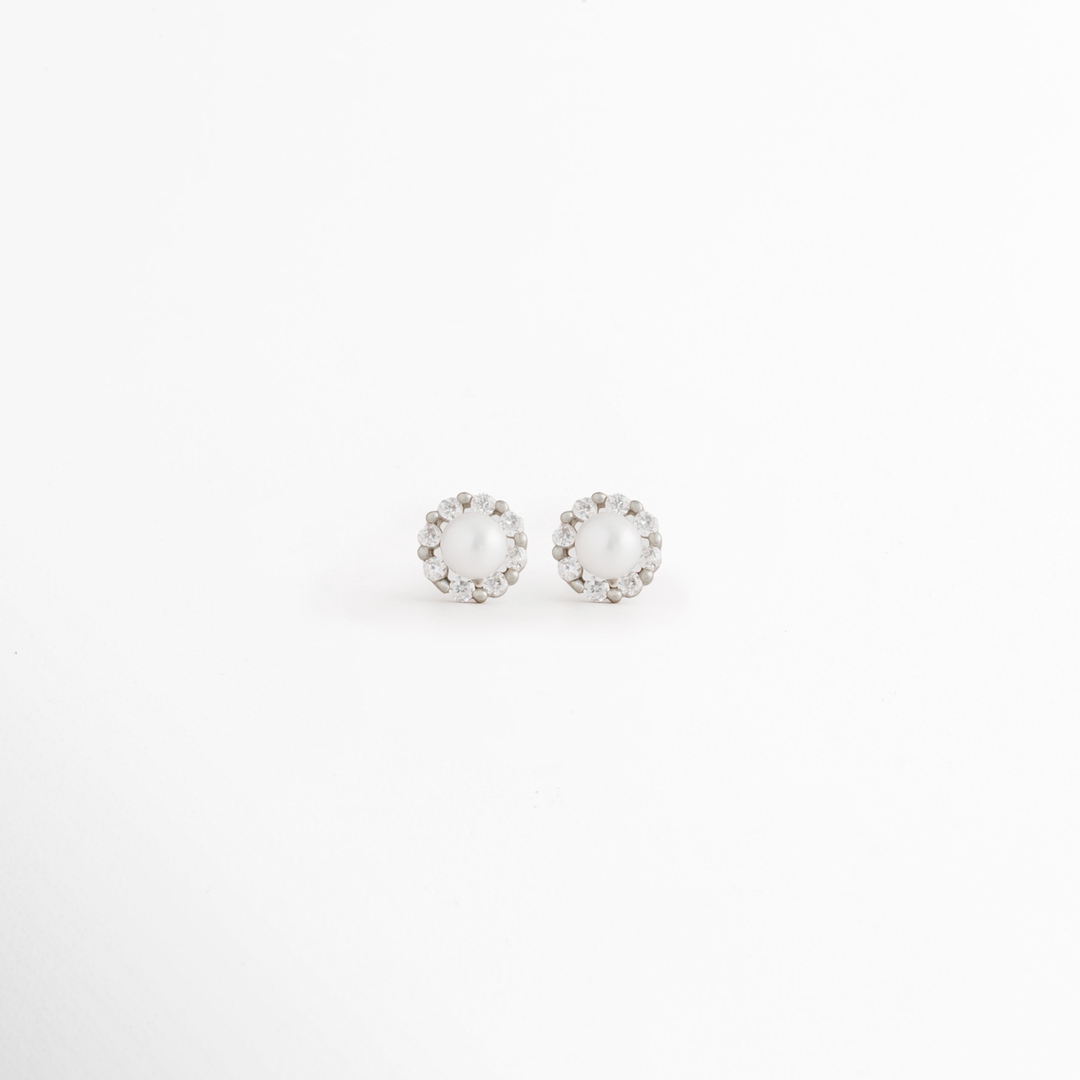 Aretes Zerina Silver Studs de Plata de Ley 925 con recubrimiento en Oro Blanco 14K y zirconia brillante sobre fondo blanco, Vista frontal de los Aretes Zerina Silver Studs con zirconia central rodeada por un delicado halo brillante, Aretes Zerina Silver Studs con diseño redondo y zirconia en acabado de Oro Blanco 14K, Par de Aretes Zerina Silver Studs elaborados en Plata de Ley 925 con zirconia y acabado plateado brillante, Aretes plateados Zerina Silver Studs con zirconia en diseño clásico y elegante.