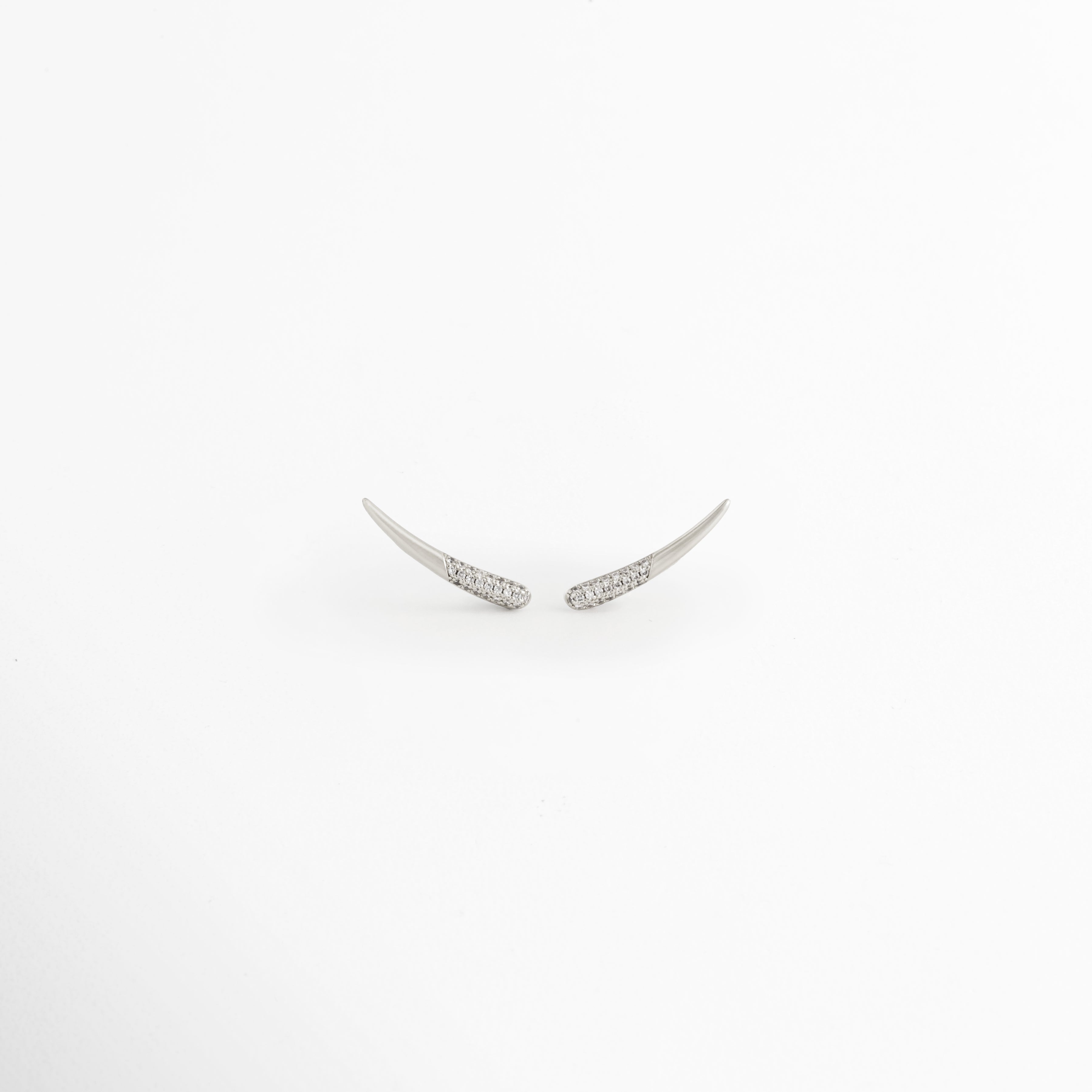 Aretes Silver Zerald de cobre con recubrimiento en Oro Blanco e incrustaciones de zirconias sobre fondo blanco, Vista frontal de los Aretes Silver Zerald con diseño curvo y zirconias brillantes, Aretes Silver Zerald con diseño minimalista curvo y acabado en Oro Blanco, Par de Aretes Silver Zerald con zirconias incrustadas y acabado plateado brillante, Aretes Silver Zerald con diseño elegante de líneas curvas y zirconias.