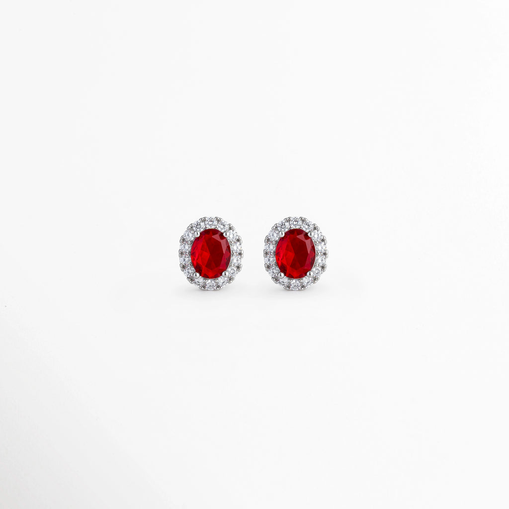 Aretes Crimson Studs enchapados en platino con circonia ovalada central en rojo rubí y halo de pedrería transparente, vista de frente sobre fondo blanco. vista frontal de aretes broquel plateados de 1.4 cm de largo con piedra roja vibrante y aureola de circonias brillantes, Plano general de aretes tipo stud de plata ley 925 platinada con gema ovalada roja estilo rubí, Crimson Studs destacando el intenso tono rojo rubí y los destellos del halo de circonias de 1.2 cm 