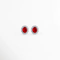 Aretes Crimson Studs enchapados en platino con circonia ovalada central en rojo rubí y halo de pedrería transparente, vista de frente sobre fondo blanco. vista frontal de aretes broquel plateados de 1.4 cm de largo con piedra roja vibrante y aureola de circonias brillantes, Plano general de aretes tipo stud de plata ley 925 platinada con gema ovalada roja estilo rubí, Crimson Studs destacando el intenso tono rojo rubí y los destellos del halo de circonias de 1.2 cm 