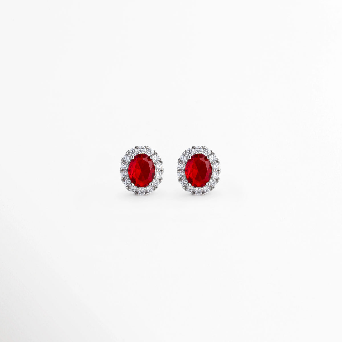 Aretes Crimson Studs enchapados en platino con circonia ovalada central en rojo rubí y halo de pedrería transparente, vista de frente sobre fondo blanco. vista frontal de aretes broquel plateados de 1.4 cm de largo con piedra roja vibrante y aureola de circonias brillantes, Plano general de aretes tipo stud de plata ley 925 platinada con gema ovalada roja estilo rubí, Crimson Studs destacando el intenso tono rojo rubí y los destellos del halo de circonias de 1.2 cm 