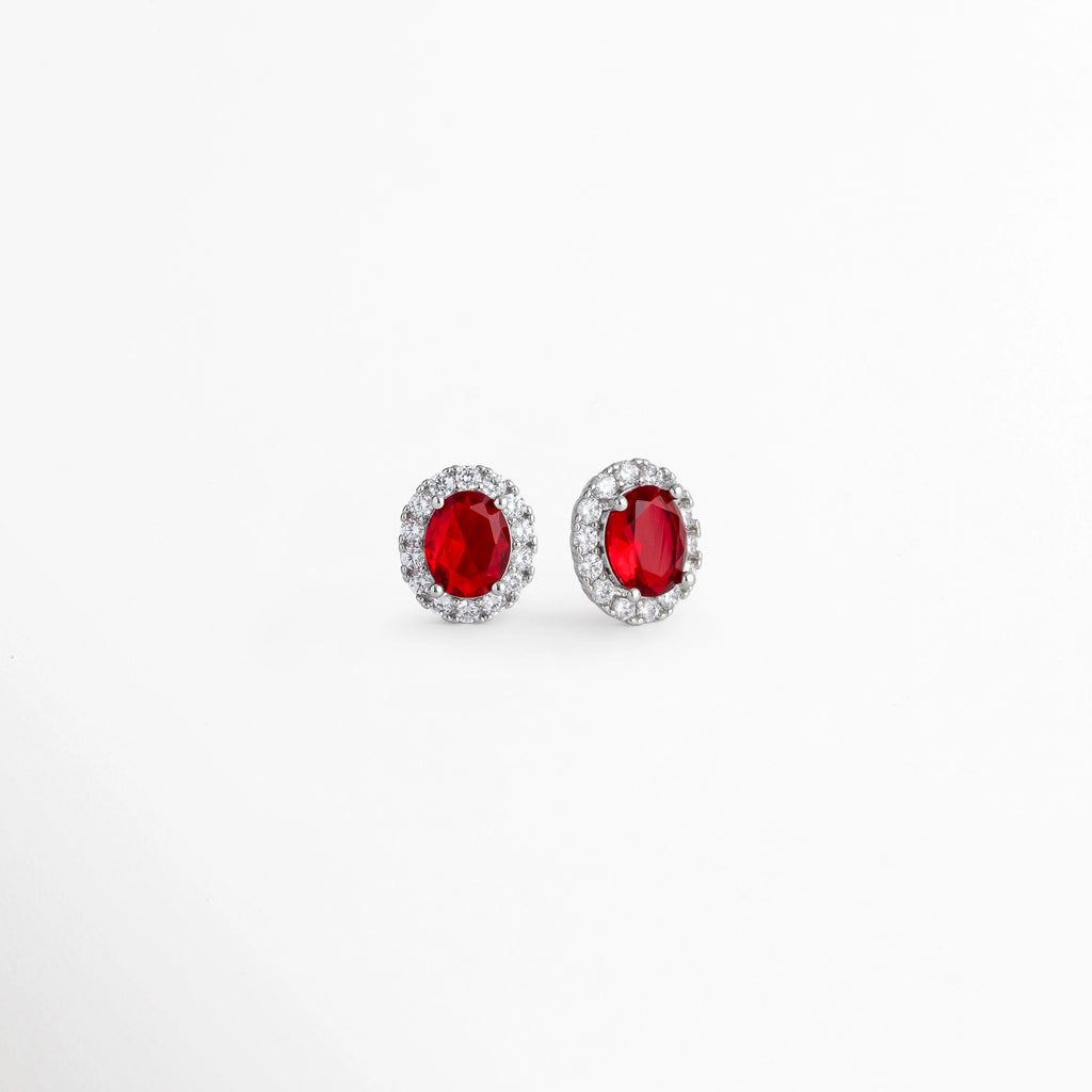 Vista en perspectiva de los aretes Crimson Studs mostrando la profundidad del engaste de la circonia roja y el borde platinado, Acercamiento en tres cuartos a los broqueles de plata ley 925 resaltando el brillo de la pedrería transparente en torno a la piedra rubí, Toma en ángulo que muestra el relieve y la corona de circonias sobre los studs de tono plata fría, Muestra de la proporción de 1.4 cm de largo y la intensidad del color carmesí de los aretes.   