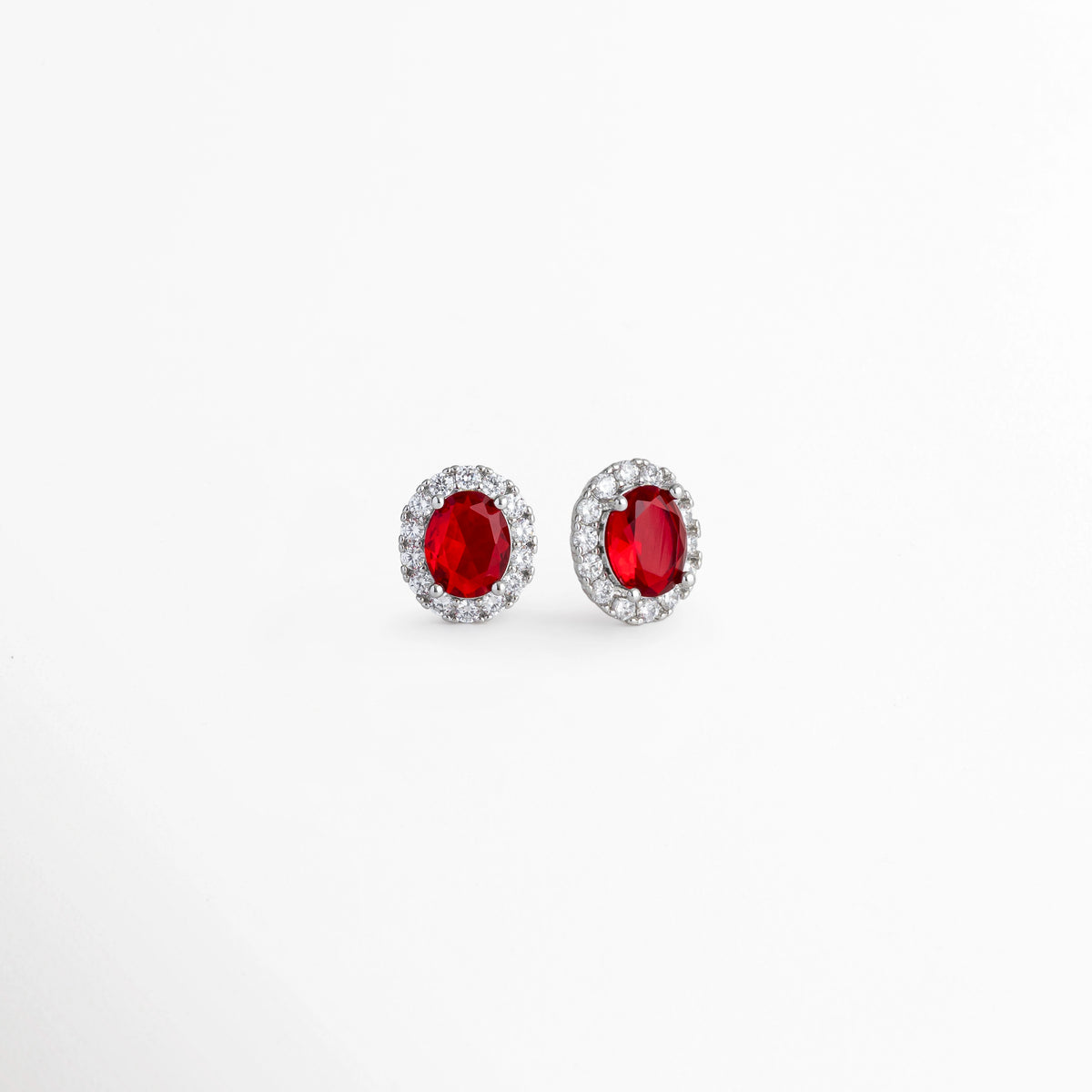 Vista en perspectiva de los aretes Crimson Studs mostrando la profundidad del engaste de la circonia roja y el borde platinado, Acercamiento en tres cuartos a los broqueles de plata ley 925 resaltando el brillo de la pedrería transparente en torno a la piedra rubí, Toma en ángulo que muestra el relieve y la corona de circonias sobre los studs de tono plata fría, Muestra de la proporción de 1.4 cm de largo y la intensidad del color carmesí de los aretes.   