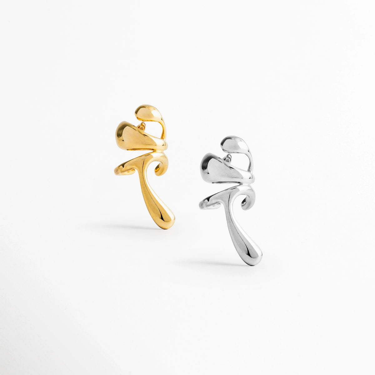 Comparativa del Ear Cuff Frago en sus dos versiones: enchapado en oro 18K y enchapado en oro blanco, Vista de par de ear cuffs de diseño orgánico en tono dorado y tono plata sobre fondo blanco, Muestra de las opciones de acabado en oro de 18K y oro blanco para el ear cuff escultural Frago, Toma conjunta de aretes de presión fluidos en colores dorado y plateado mostrando sus destellos brillantes.