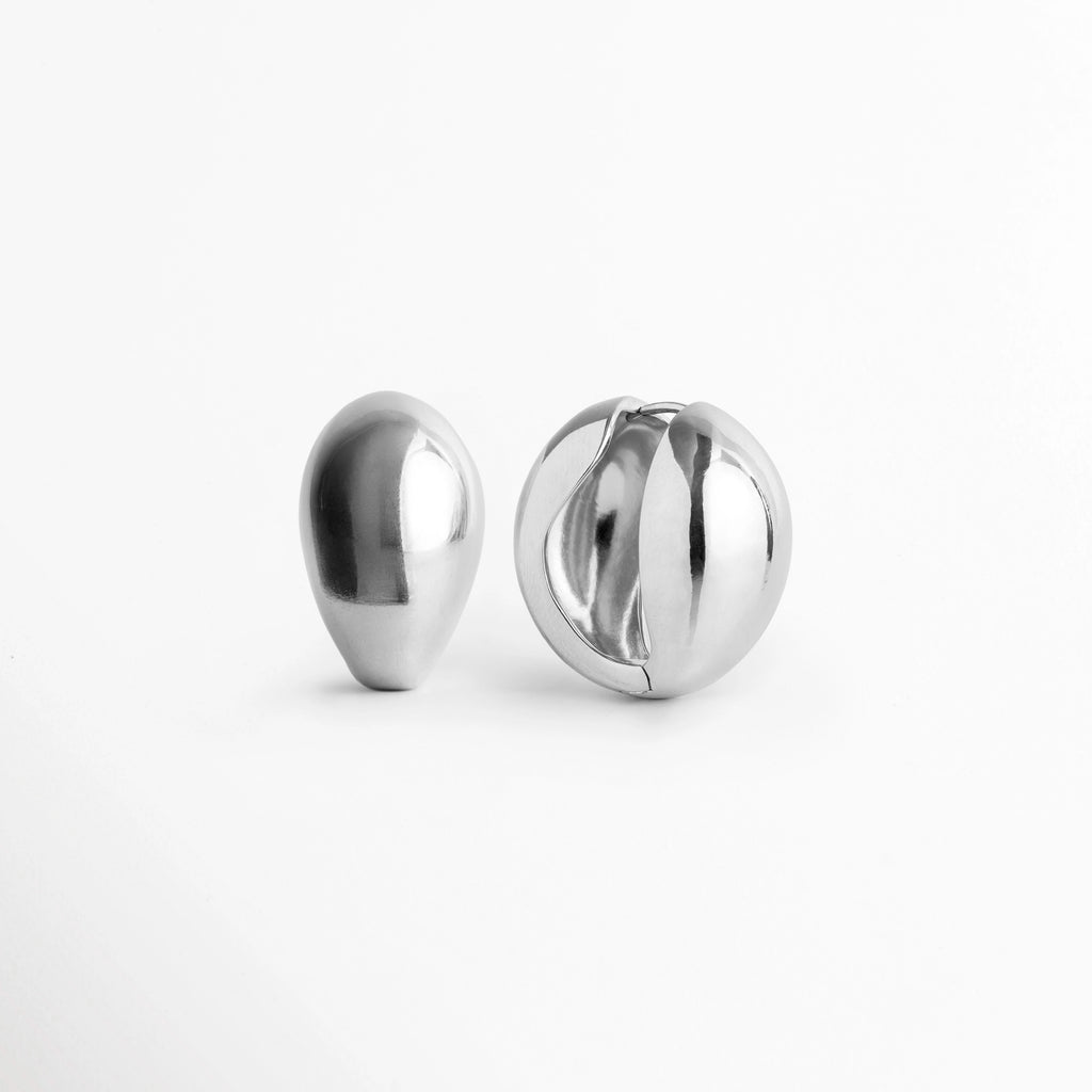Aretes Silver Nevara vistos de frente y perfil mostrando la silueta de gota de agua y la apertura central de bisagra, Vista en ángulo de arete abombado plateado con diseño cóncavo posterior y cierre articulado, Acercamiento a la silueta orgánica y al reverso hueco de los aretes en forma de gota plateada, Detalle del volumen abultado de 1.8 cm de ancho del arete enchapado en tono plata sobre superficie clara.