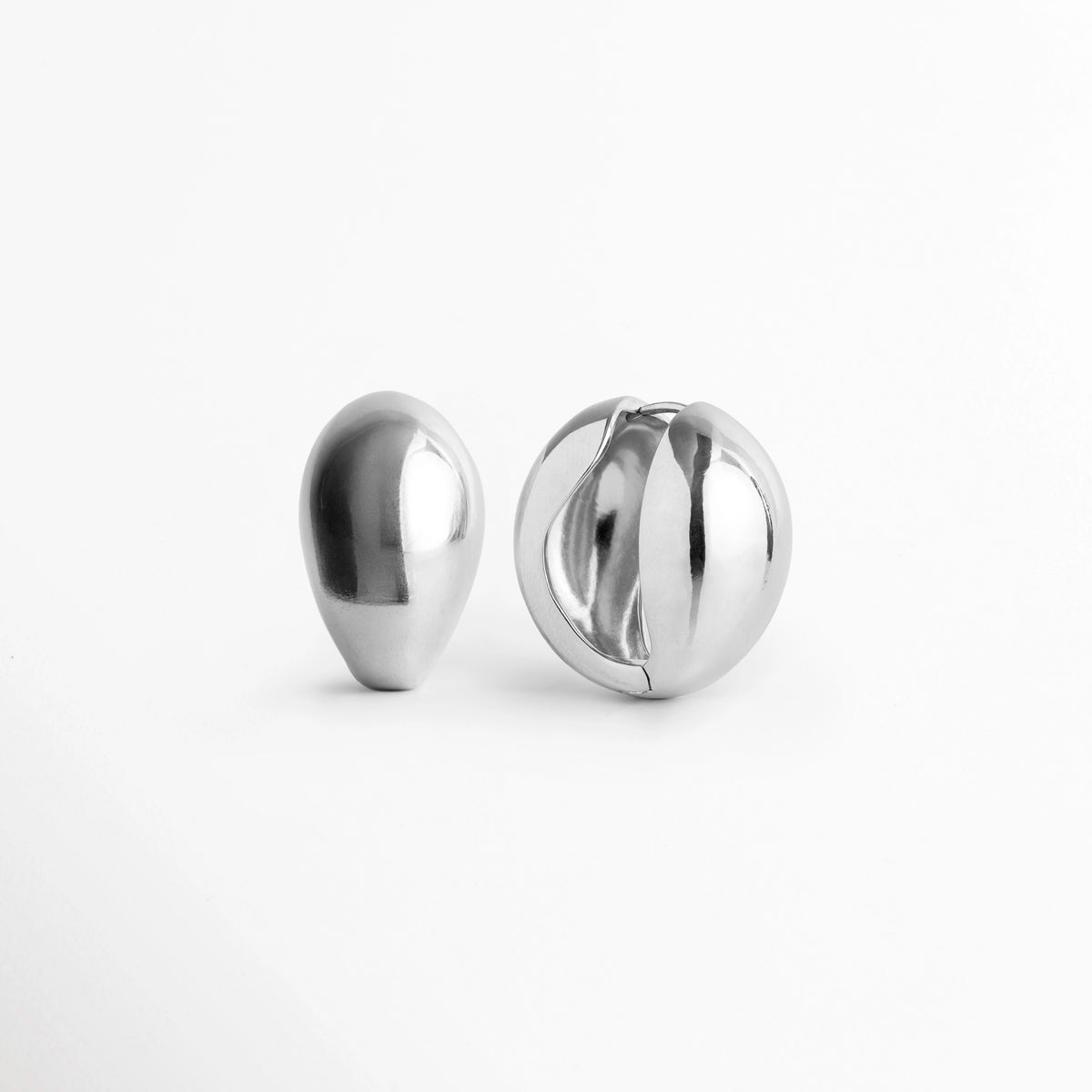 Aretes Silver Nevara vistos de frente y perfil mostrando la silueta de gota de agua y la apertura central de bisagra, Vista en ángulo de arete abombado plateado con diseño cóncavo posterior y cierre articulado, Acercamiento a la silueta orgánica y al reverso hueco de los aretes en forma de gota plateada, Detalle del volumen abultado de 1.8 cm de ancho del arete enchapado en tono plata sobre superficie clara.