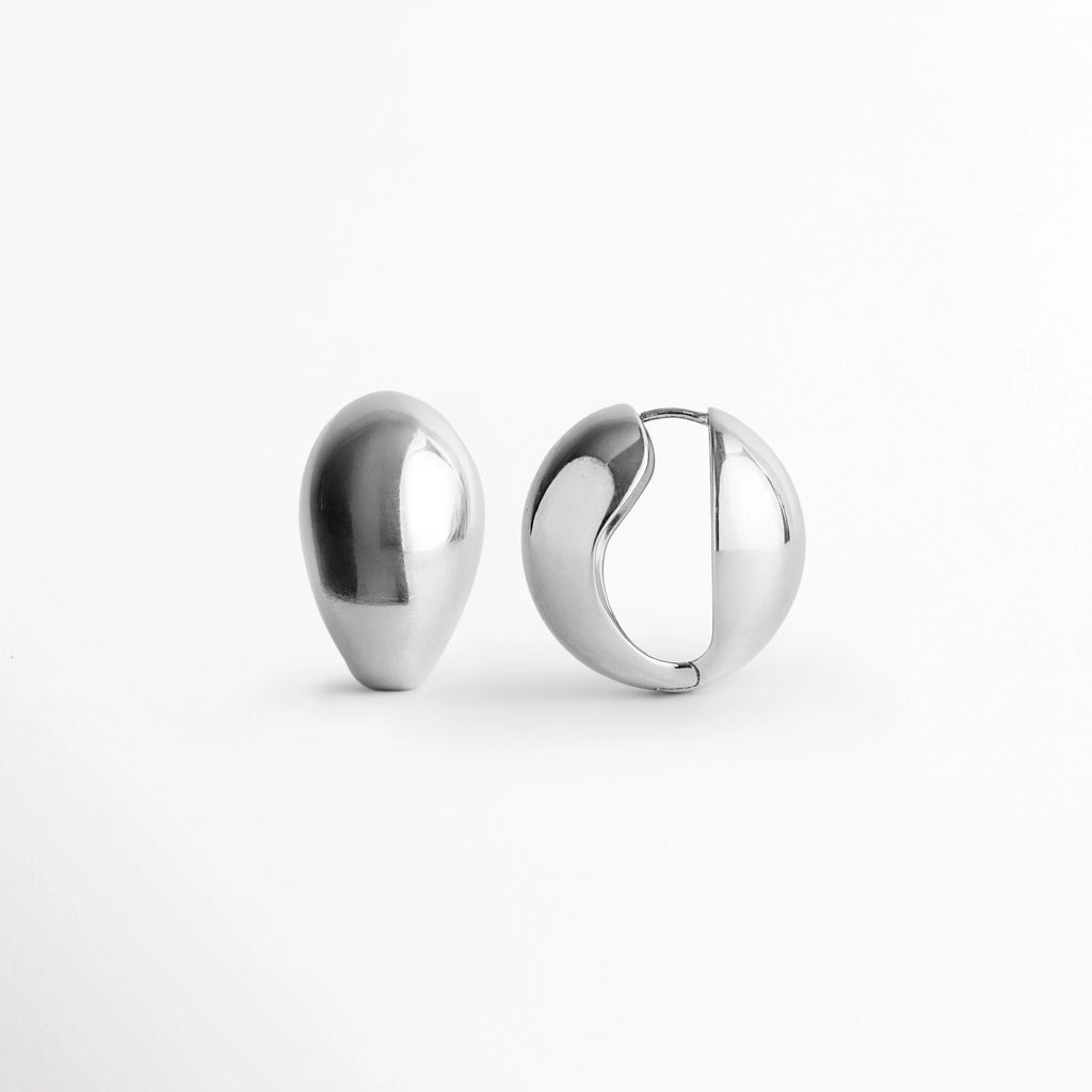 Arete Silver Nevara de perfil con la bisagra ligeramente abierta mostrando el poste integrado, Vista lateral del sistema de cierre snap/huggie de la arracada abombada plateada en forma de gota, Acercamiento al mecanismo de apertura en bisagra de los aretes plateados de volumen francés, Detalle del poste curvo y el encaje de seguridad de la arracada en forma de gota con acabado brillante.