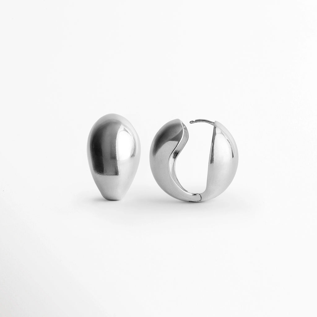 Vista lateral de los Aretes Silver Nevara mostrando la articulación del aro totalmente abierta para colocación, Detalle del broche de encaje oculto y la estructura interna liviana del arete plateado en forma de gota, Muestra del mecanismo de fácil uso y ajuste seguro en el arete abombado con acabado plateado espejo, Vista de cerca de la bisagra articulada de los aretes de volumen fluido sobre fondo claro.