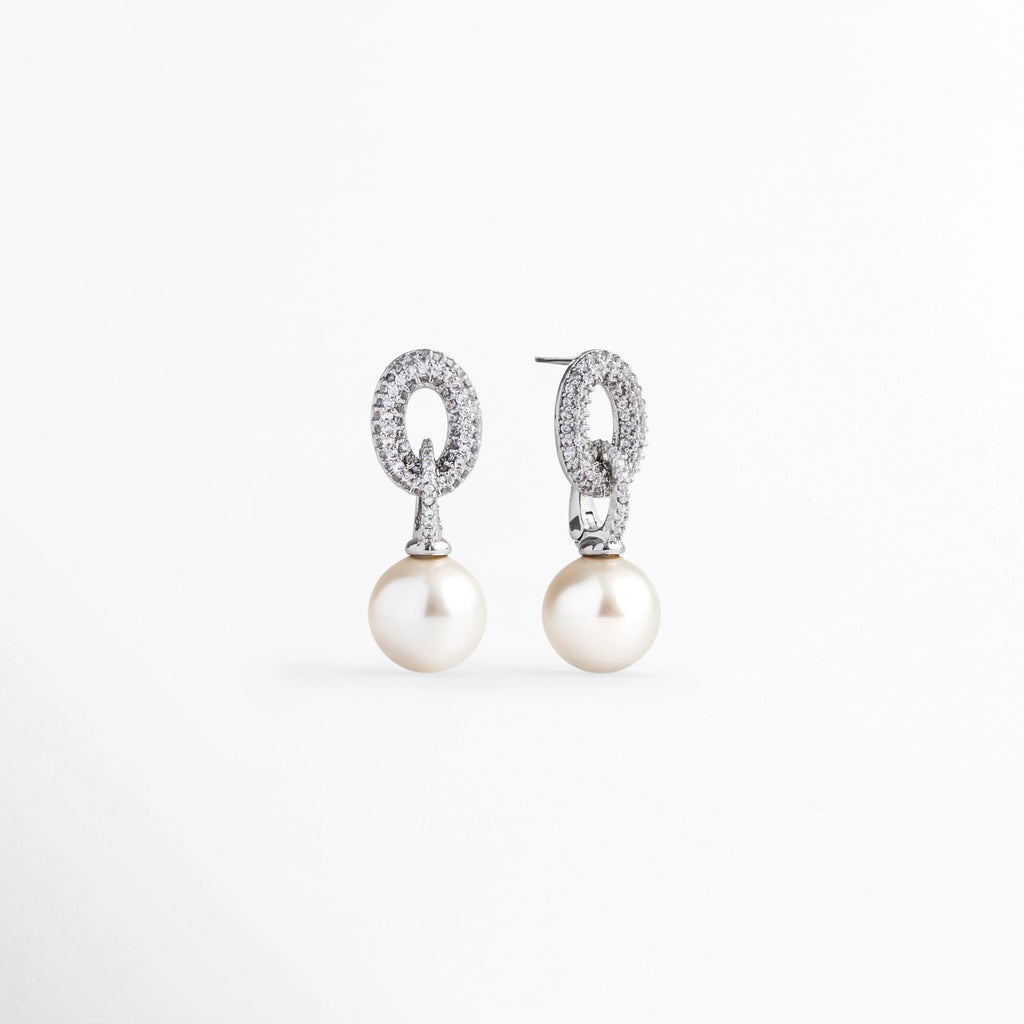Vista en ángulo de los aretes de perla mostrando el volumen de la gota esférica y el destello del eslabón superior de circonias, Acercamiento en perspectiva al soporte metálico plateado cuajado de circonias con perla colgante de 2.7 cm, Toma en diagonal que destaca el contraste entre el brillo cristalino de las circonias y el lustre nacarado de la perla, Muestra de la caída fluida y la proporción delicada de 1 cm de ancho de los aretes de perla sintética.
