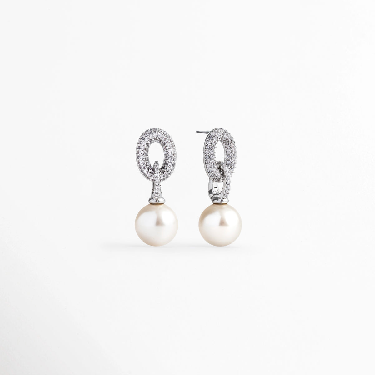 Vista en ángulo de los aretes de perla mostrando el volumen de la gota esférica y el destello del eslabón superior de circonias, Acercamiento en perspectiva al soporte metálico plateado cuajado de circonias con perla colgante de 2.7 cm, Toma en diagonal que destaca el contraste entre el brillo cristalino de las circonias y el lustre nacarado de la perla, Muestra de la caída fluida y la proporción delicada de 1 cm de ancho de los aretes de perla sintética.