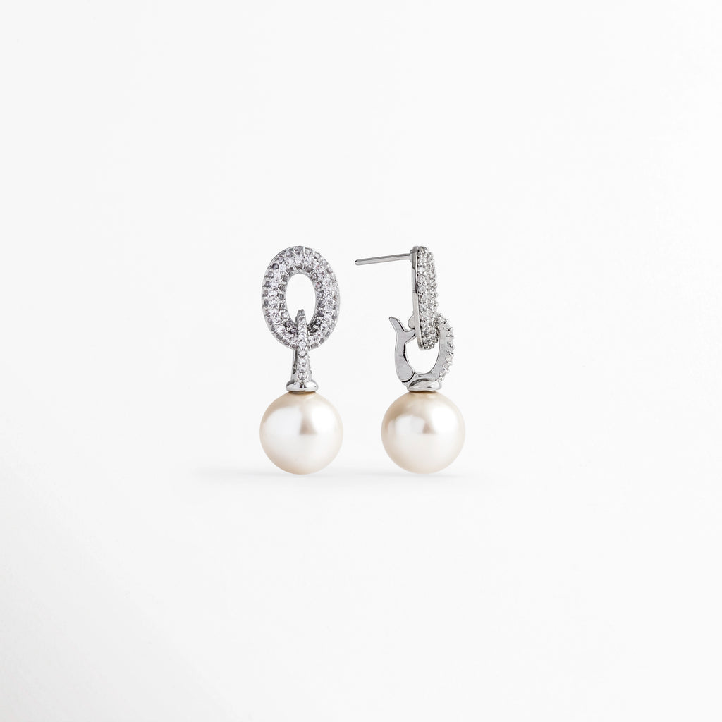 Vista de perfil de los aretes de perla mostrando el poste recto de ajuste para oreja perforada, Detalle del reverso pulido y sistema de sujeción de poste en el eslabón de circonias de los aretes, Acercamiento lateral a la montura plateada enchapada y el acabado nacarado de la perla de 1 cm, Muestra de perfil de la estructura ligera de 2.7 cm de longitud con cierre seguro.
