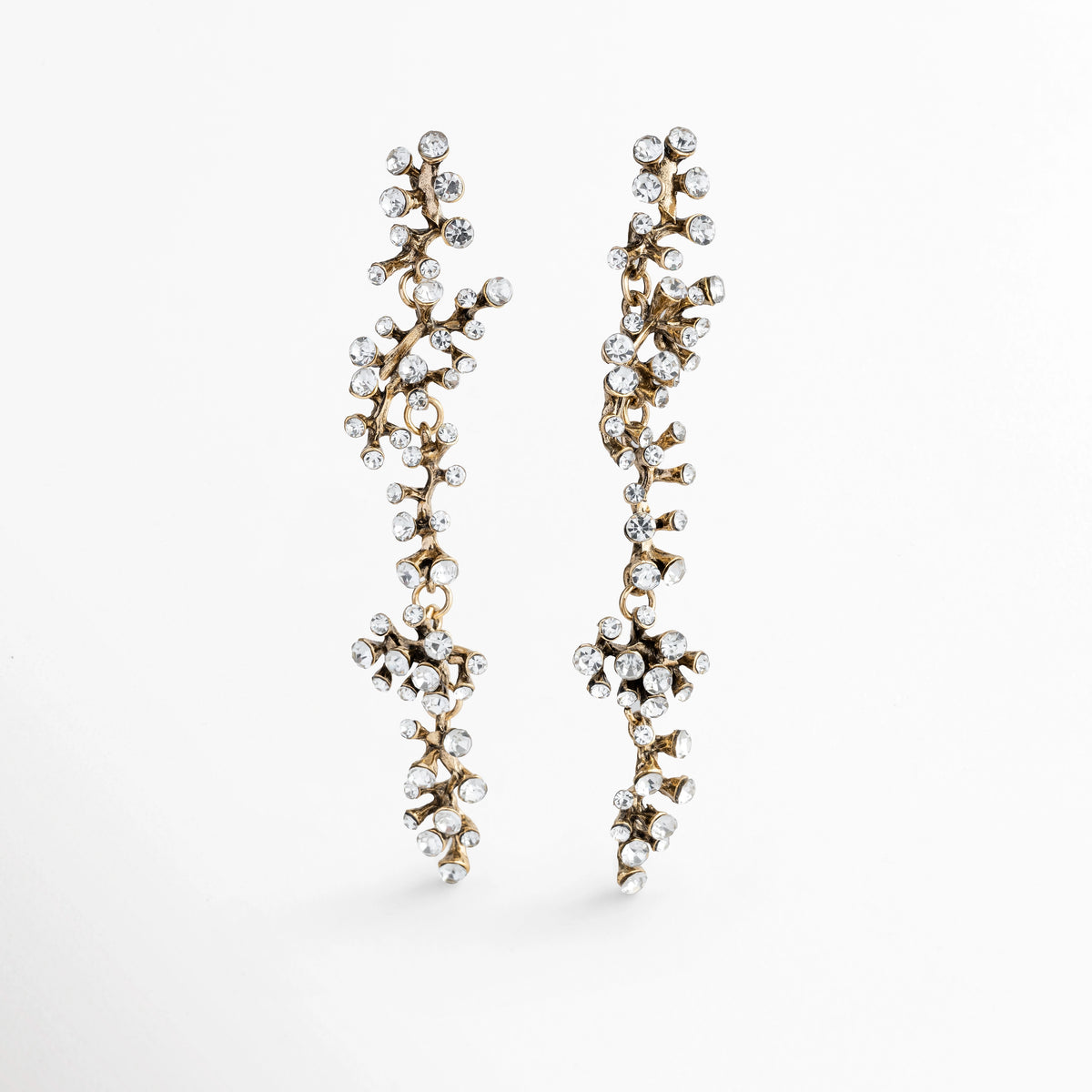 Vista en ángulo de los Aretes Laurel mostrando el relieve tridimensional de sus ramas con circonias e incrustaciones, Acercamiento en perspectiva de aretes largos de 9.5 cm con textura botánica de laurel y gemas transparentes, Toma en diagonal que destaca el ancho de 1.7 cm de las ramificaciones y el brillo de la pedrería, Muestra de la caída estilizada y ligereza visual de los aretes largos de acero inoxidable con circonias.