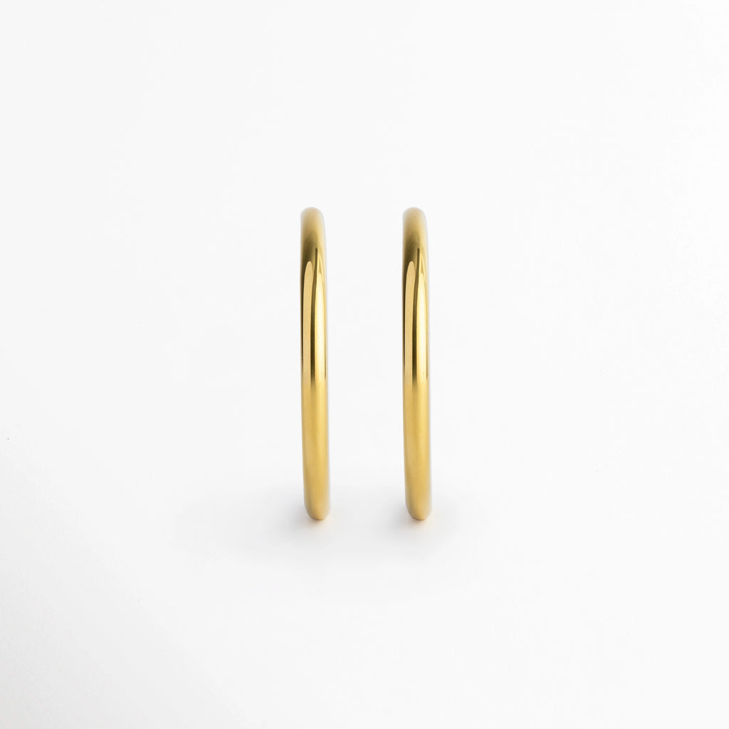 Vista de frente de los Aretes Adele mostrando el grosor uniforme de 0.4 cm de la tubuladura metálica, Par de arracadas doradas en posición vertical paralela evidenciando su acabado pulido brillante, Muestra del perfil frontal ultra estilizado y simétrico de los aros enchapados en oro 18K, Vista frontal minimalista del grosor tubular de 0.4 cm de las arracadas en acero inoxidable.