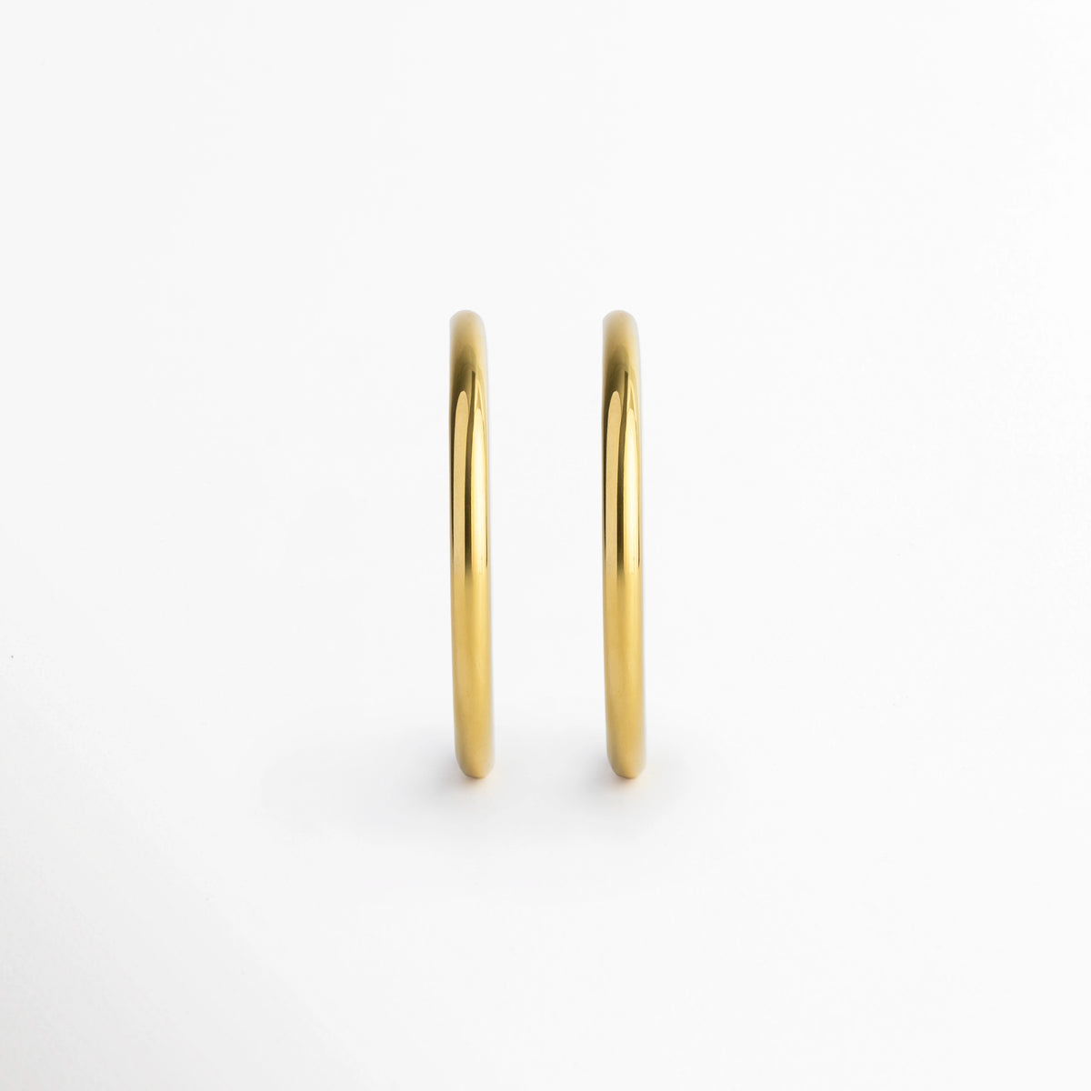 Vista de frente de los Aretes Adele mostrando el grosor uniforme de 0.4 cm de la tubuladura metálica, Par de arracadas doradas en posición vertical paralela evidenciando su acabado pulido brillante, Muestra del perfil frontal ultra estilizado y simétrico de los aros enchapados en oro 18K, Vista frontal minimalista del grosor tubular de 0.4 cm de las arracadas en acero inoxidable.