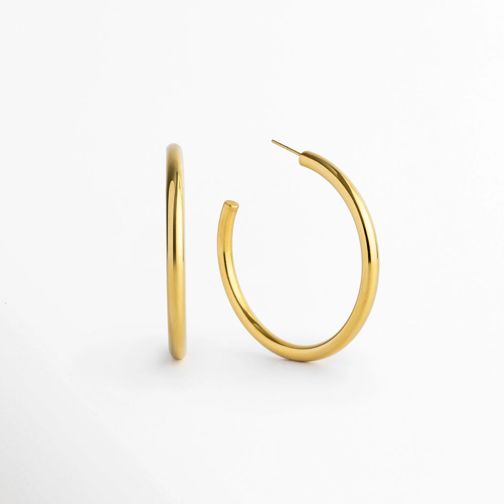 Aretes Adele dorados de 5 cm de diámetro con tubo de 0.4 cm de grosor enchapados en oro 18K, vista en ángulo sobre fondo blanco, Vista frontal y lateral de par de arracadas de aro mediano-grande en acero inoxidable dorado de silueta tubular, Arracadas doradas de 5 cm de diámetro con poste de ajuste e interiores huecos ligeros en oro 18K, Par de aros dorados clásicos de perfil circular continuo de 0.4 cm de grosor para uso diario o de noche.