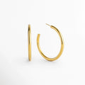 Aretes Adele dorados de 5 cm de diámetro con tubo de 0.4 cm de grosor enchapados en oro 18K, vista en ángulo sobre fondo blanco, Vista frontal y lateral de par de arracadas de aro mediano-grande en acero inoxidable dorado de silueta tubular, Arracadas doradas de 5 cm de diámetro con poste de ajuste e interiores huecos ligeros en oro 18K, Par de aros dorados clásicos de perfil circular continuo de 0.4 cm de grosor para uso diario o de noche.