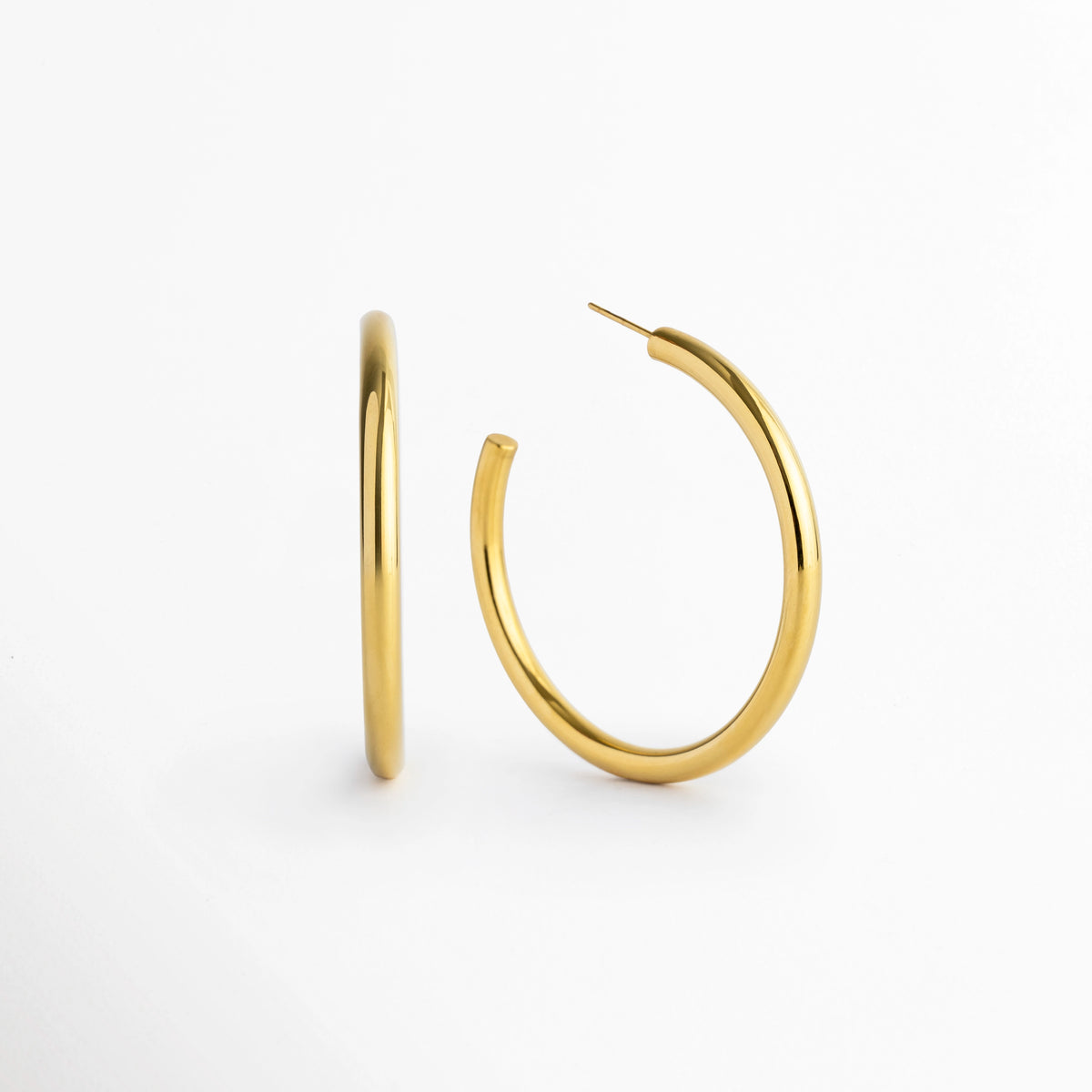 Aretes Adele dorados de 5 cm de diámetro con tubo de 0.4 cm de grosor enchapados en oro 18K, vista en ángulo sobre fondo blanco, Vista frontal y lateral de par de arracadas de aro mediano-grande en acero inoxidable dorado de silueta tubular, Arracadas doradas de 5 cm de diámetro con poste de ajuste e interiores huecos ligeros en oro 18K, Par de aros dorados clásicos de perfil circular continuo de 0.4 cm de grosor para uso diario o de noche.