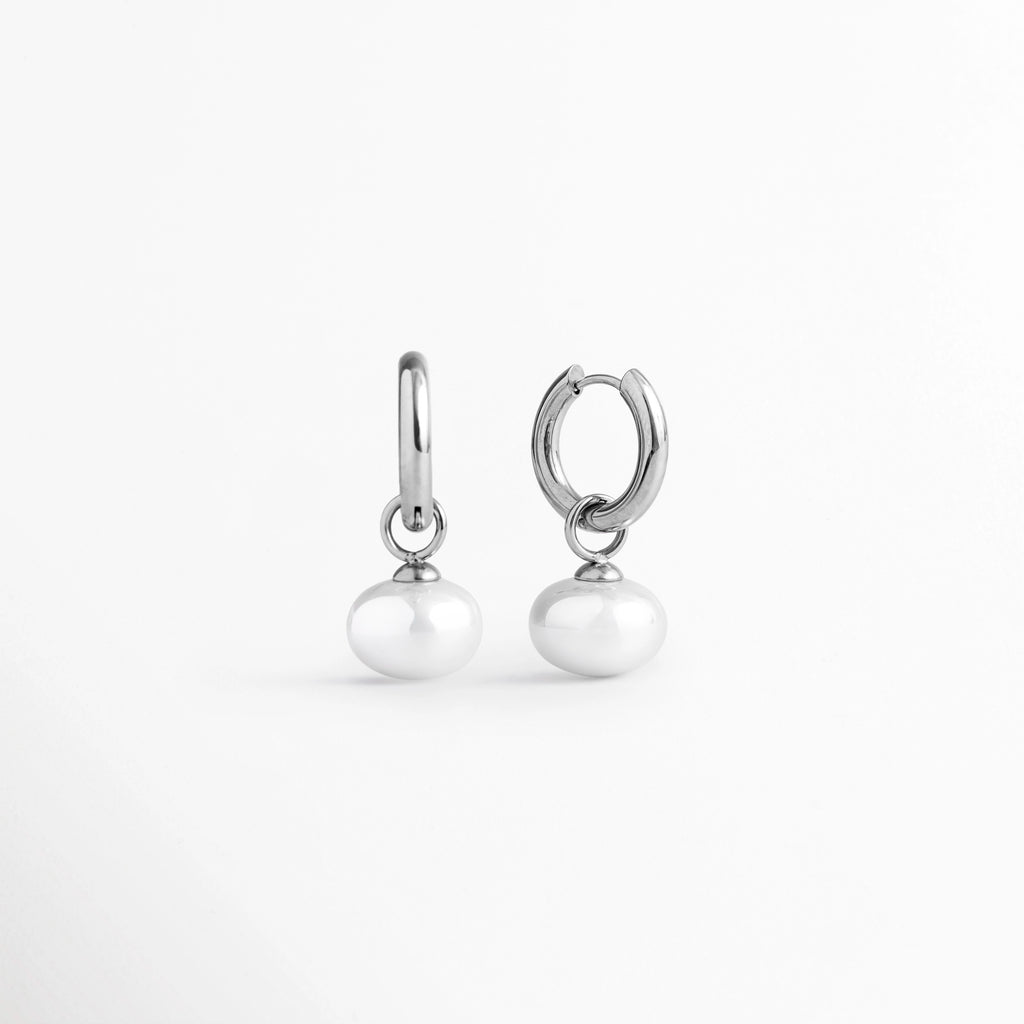 Vista de perfil de las arracadas Dune Silver Hoops mostrando la curvatura del aro plateado y el dije de perla sintética, Acercamiento lateral a los aretes huggie plateados resaltando el brillo del enchapado en oro blanco 16K y el tono nacarado de la perla, Toma en ángulo que muestra la caída natural de la perla suspendida en el aro de acero inoxidable plateado, Muestra de la proporción de 3 cm de largo del arete plateado y el ensamble de la perla sintética.