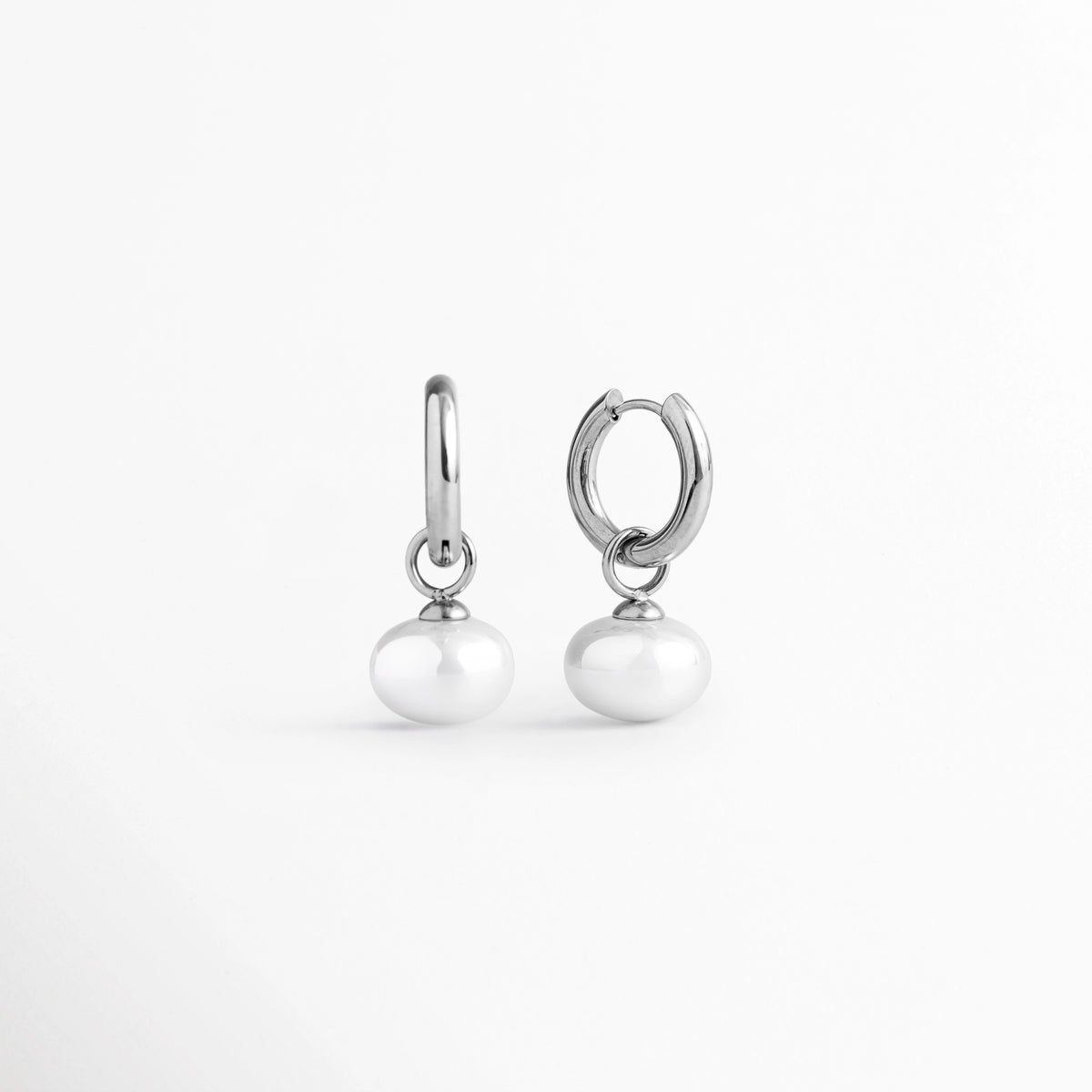 Vista de perfil de las arracadas Dune Silver Hoops mostrando la curvatura del aro plateado y el dije de perla sintética, Acercamiento lateral a los aretes huggie plateados resaltando el brillo del enchapado en oro blanco 16K y el tono nacarado de la perla, Toma en ángulo que muestra la caída natural de la perla suspendida en el aro de acero inoxidable plateado, Muestra de la proporción de 3 cm de largo del arete plateado y el ensamble de la perla sintética.