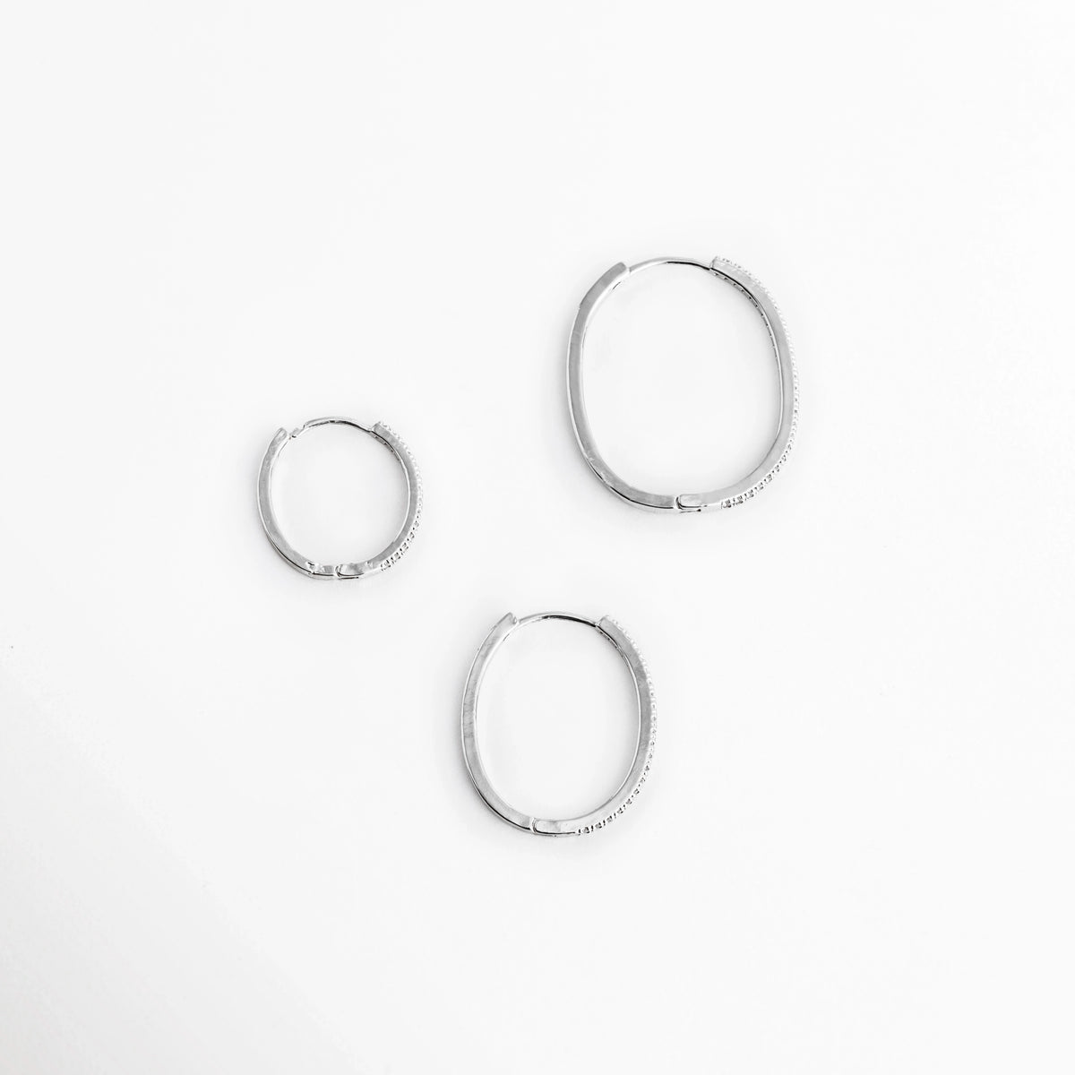 Muestra de los tres tamaños individuales de arracadas plateadas (2.1 cm, 2.9 cm y 3.2 cm) distribuidos sobre superficie blanca, Comparación de escala de los tres aros delgados plateados con pavé de circonias transparentes, Vista superior de tres arracadas con acabado plateado ordenadas por dimensión de menor a mayor, Trío de aros estilo huggie y arracada media plateados con incrustaciones de circonias cristalinas.