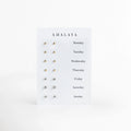 Set Day by Day de Amalaya compuesto por siete pares de aretes broquel enchapados en oro 14k presentados en tarjeta con los días de la semana, vista frontal sobre fondo blanco, Vista de frente del juego de 7 pares de aretes stud en tono oro de 14k, incluyendo cuatro pares de esferas pulidas y tres pares de circonias transparentes engastadas, Plano general de set semanal de broqueles dorados para cada día de la semana, desde Monday hasta Sunday, sobre tarjeta de exhibición blanca. 