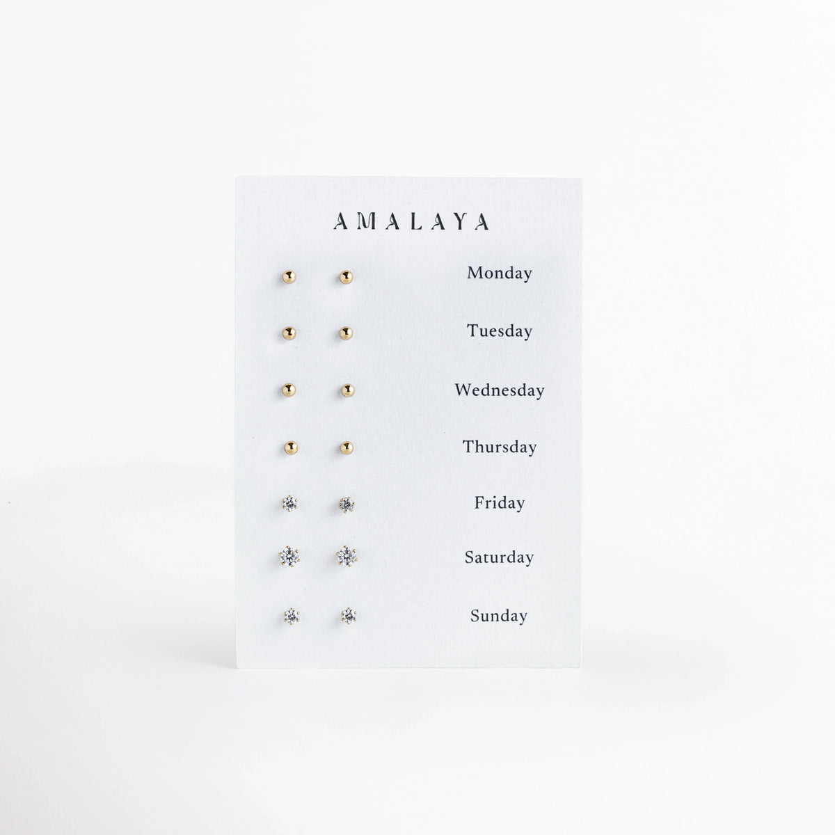 Set Day by Day de Amalaya compuesto por siete pares de aretes broquel enchapados en oro 14k presentados en tarjeta con los días de la semana, vista frontal sobre fondo blanco, Vista de frente del juego de 7 pares de aretes stud en tono oro de 14k, incluyendo cuatro pares de esferas pulidas y tres pares de circonias transparentes engastadas, Plano general de set semanal de broqueles dorados para cada día de la semana, desde Monday hasta Sunday, sobre tarjeta de exhibición blanca. 