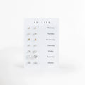 Set All Week de Amalaya compuesto por siete pares de aretes broquel enchapados en oro 14k de distintos tamaños presentados en tarjeta semanal, vista frontal sobre fondo blanco, Vista de frente del juego semanal de 7 pares de aretes stud en tono oro de 14k, incluyendo dos pares de esferas pulidas y cinco pares de circonias cristalinas de corte solitario, Plano general de set de broqueles dorados para cada día de la semana, de Monday a Sunday, ordenados por escala de tamaño