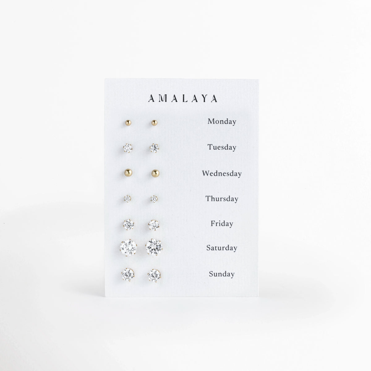 Set All Week de Amalaya compuesto por siete pares de aretes broquel enchapados en oro 14k de distintos tamaños presentados en tarjeta semanal, vista frontal sobre fondo blanco, Vista de frente del juego semanal de 7 pares de aretes stud en tono oro de 14k, incluyendo dos pares de esferas pulidas y cinco pares de circonias cristalinas de corte solitario, Plano general de set de broqueles dorados para cada día de la semana, de Monday a Sunday, ordenados por escala de tamaño